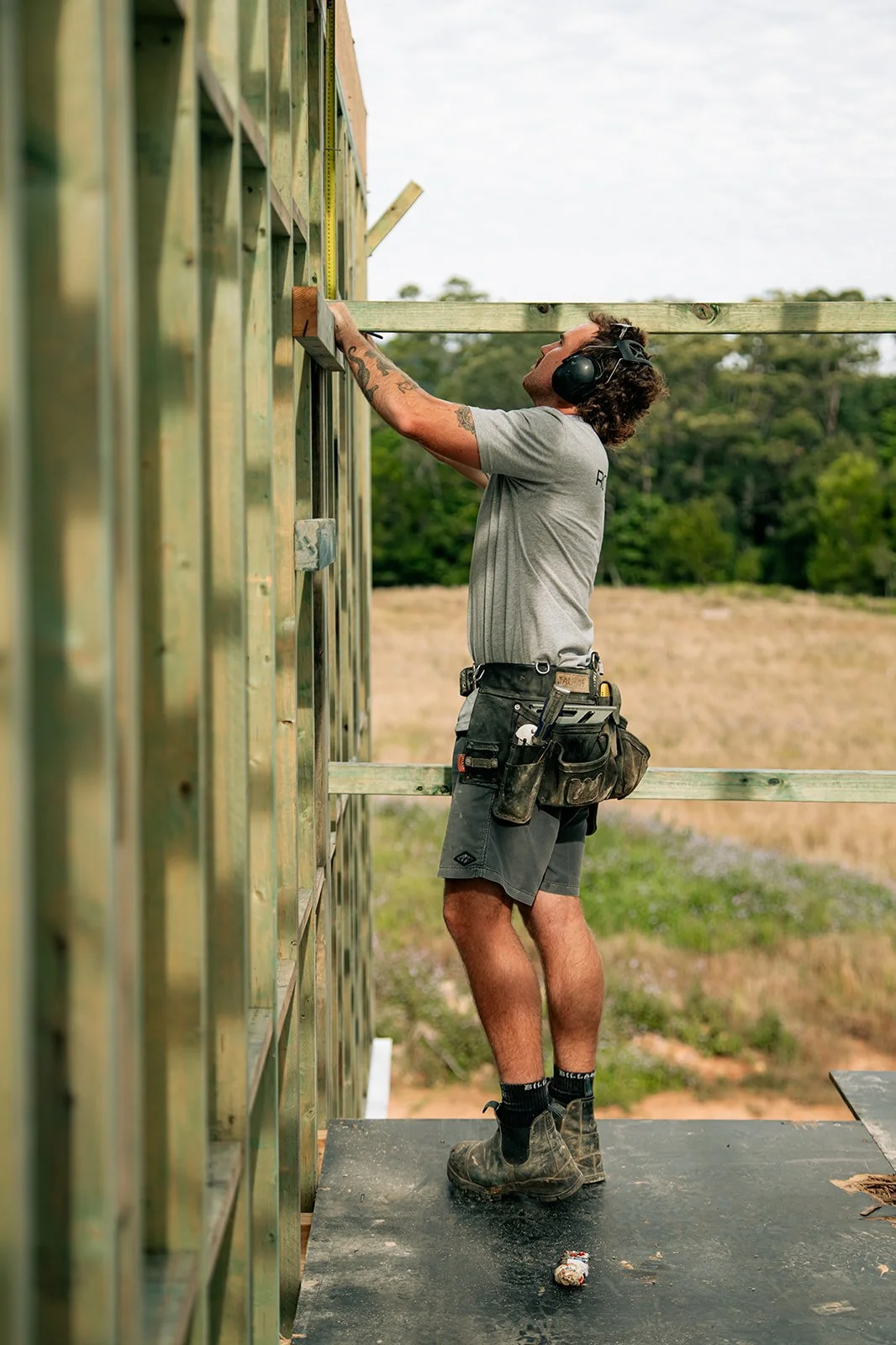 A construction worker measuring a wall at an outdoor building site, wearing headphones, shorts, safety boots, and a tool belt.