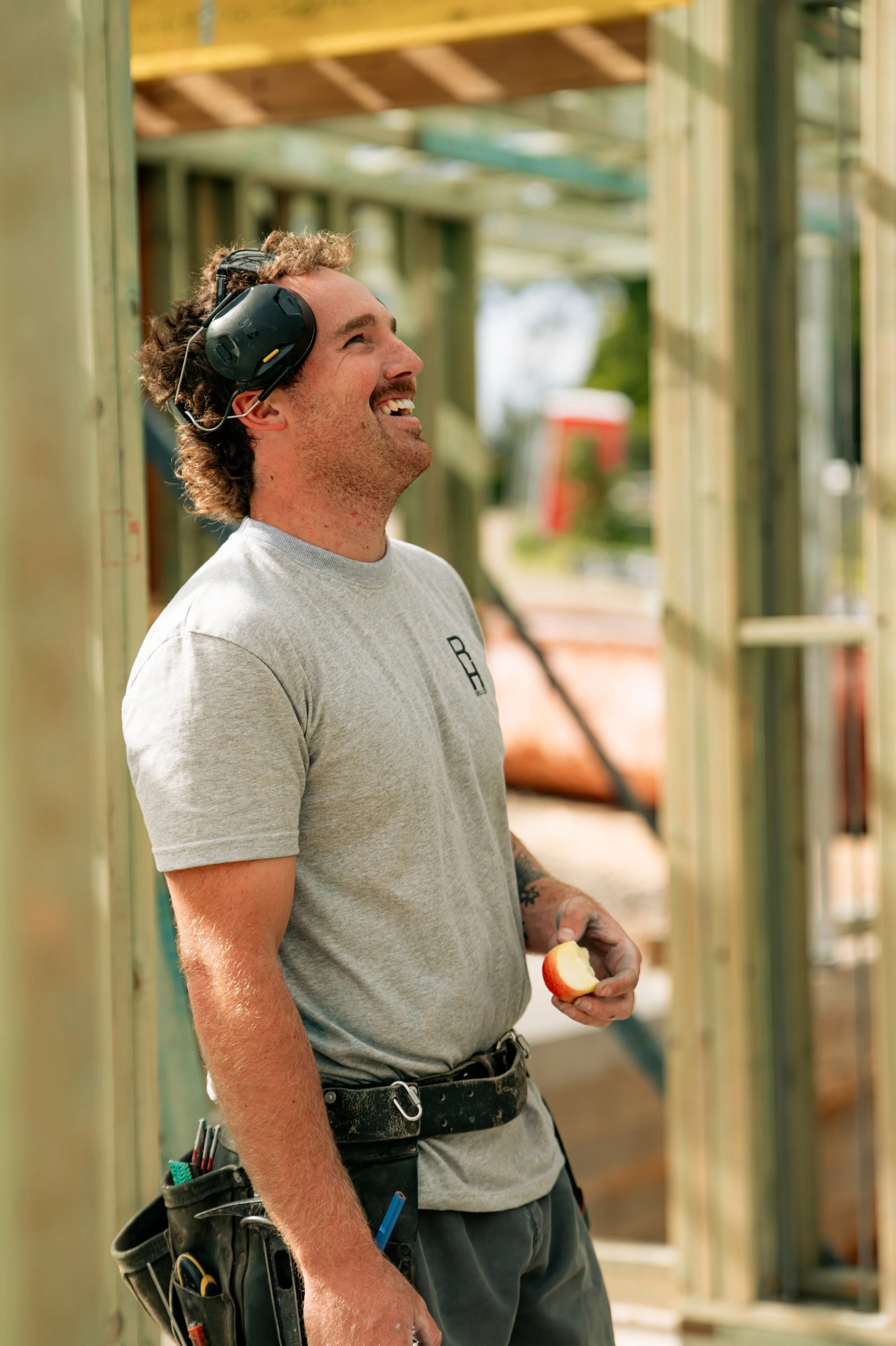 A man wearing a gray t-shirt with a tool belt, holding an apple slice, smiling while working on a building construction site.
