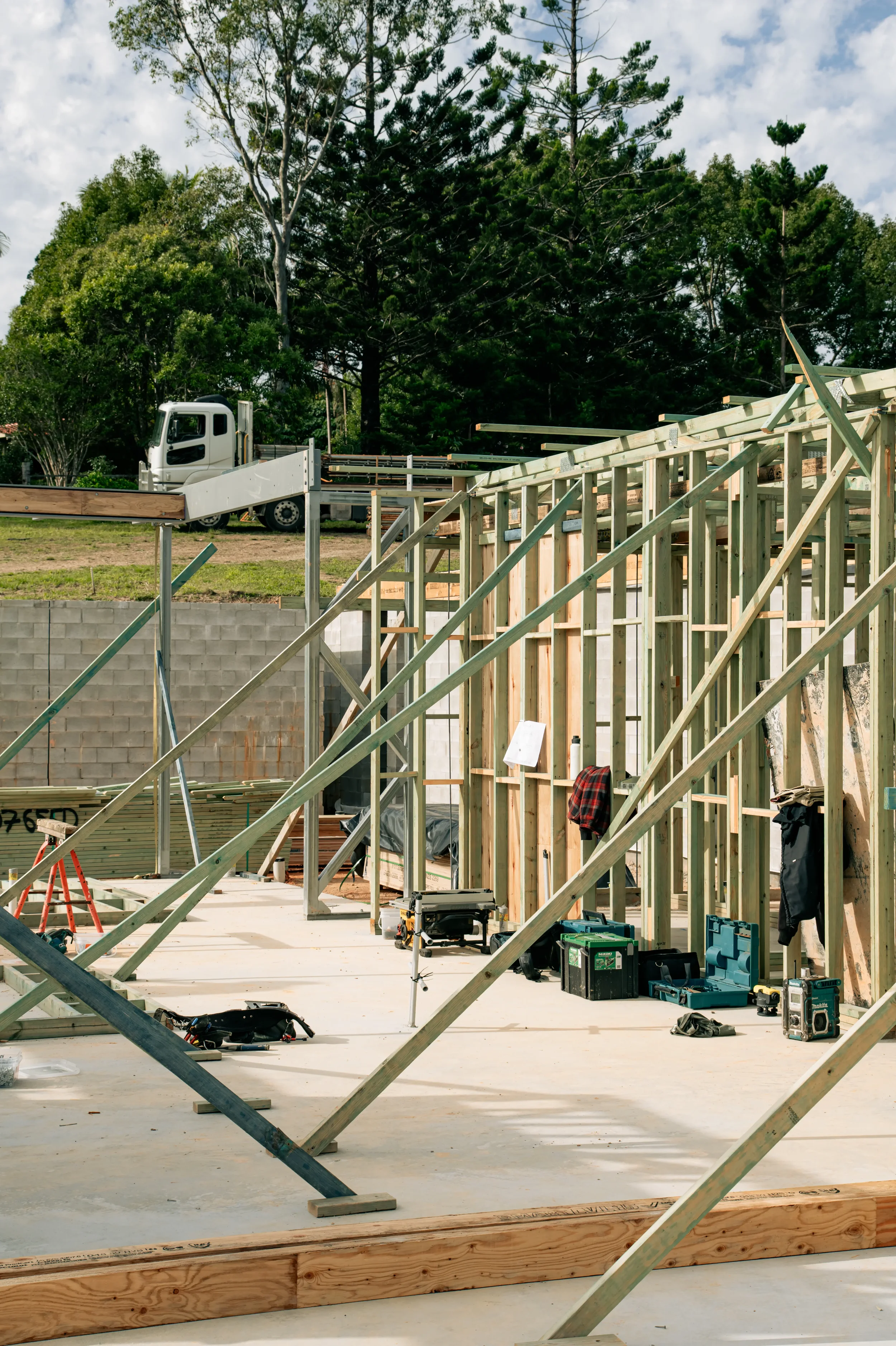 Construction site with wooden framework and tools, a white truck in the background, trees and cloudy sky.