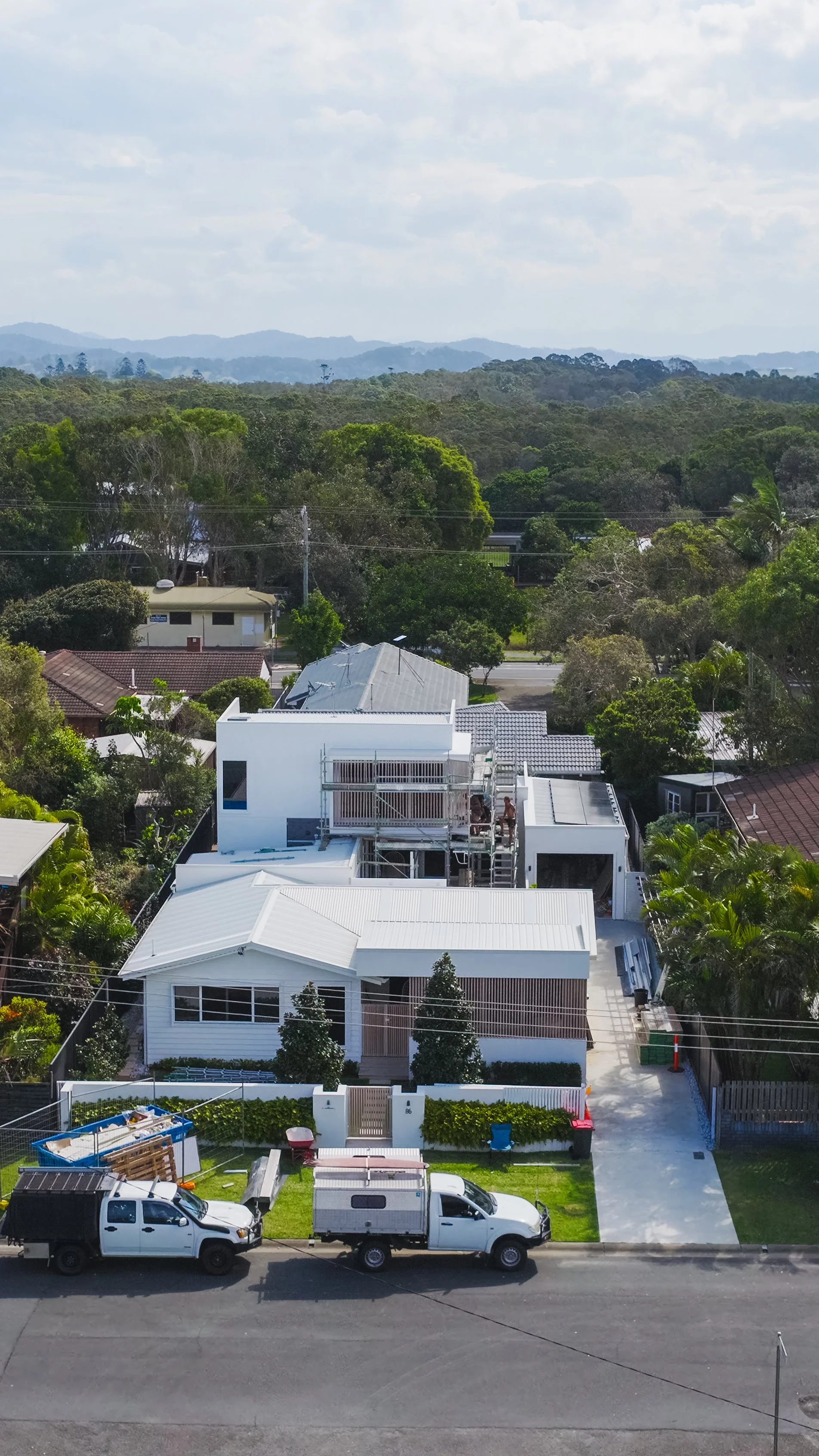 Aerial view of a residential neighborhood with a new white house under construction, surrounded by trees, other houses, and mountains in the distance.
