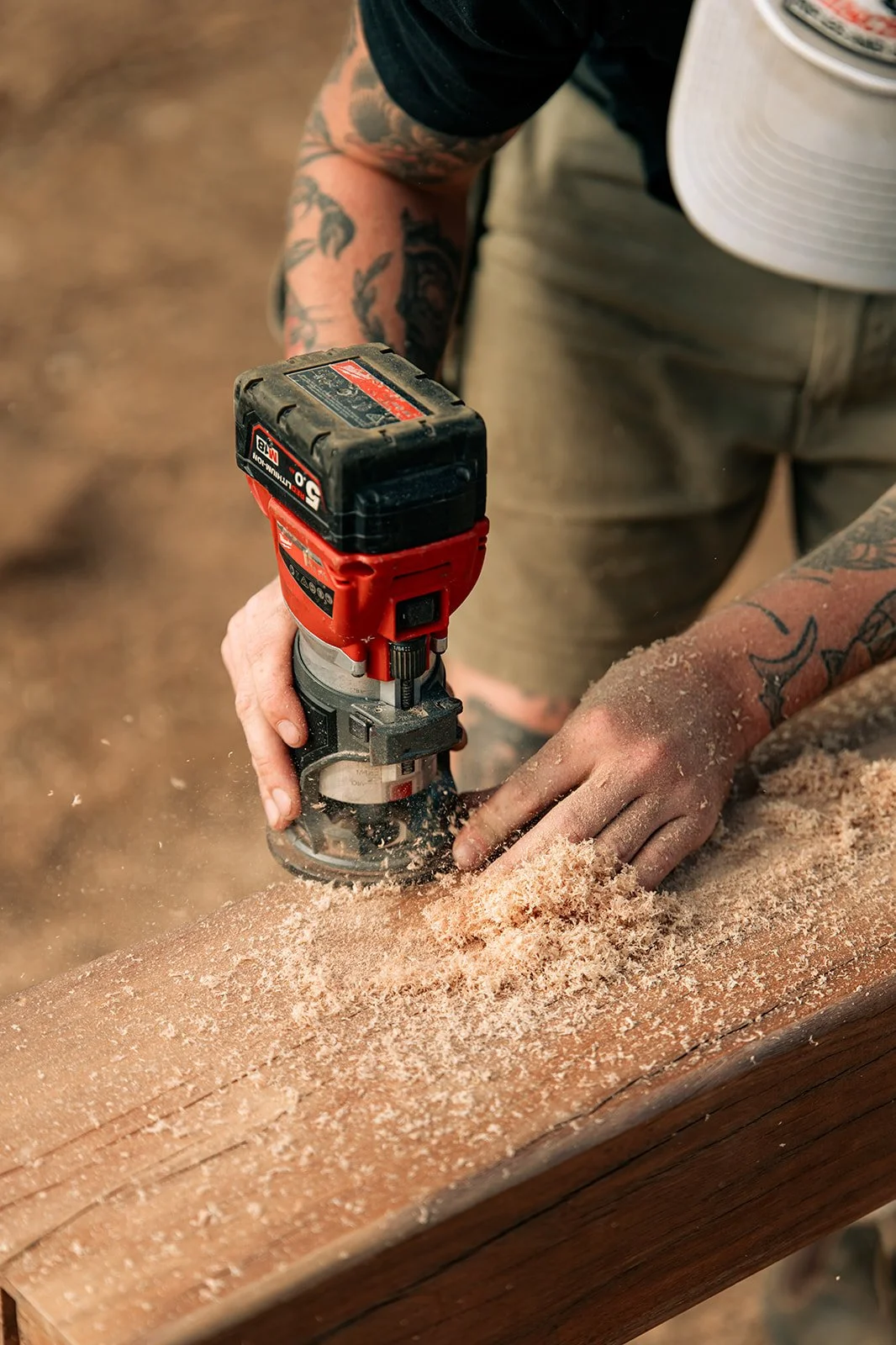 A person with tattoos is using a cordless multi-tool to sand a piece of wood, with sawdust flying around in the workshop.