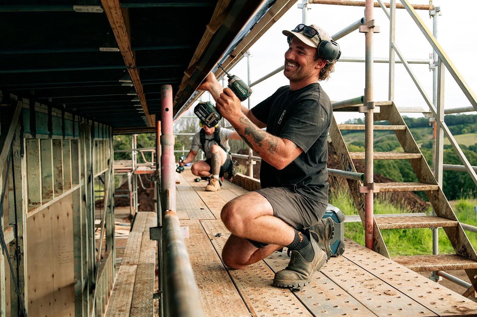 Two construction workers building a wooden structure outdoors. One worker, kneeling and smiling, is using a cordless drill, while the other, sitting nearby, is working with other tools. Scaffolding surrounds them, with green fields in the background.
