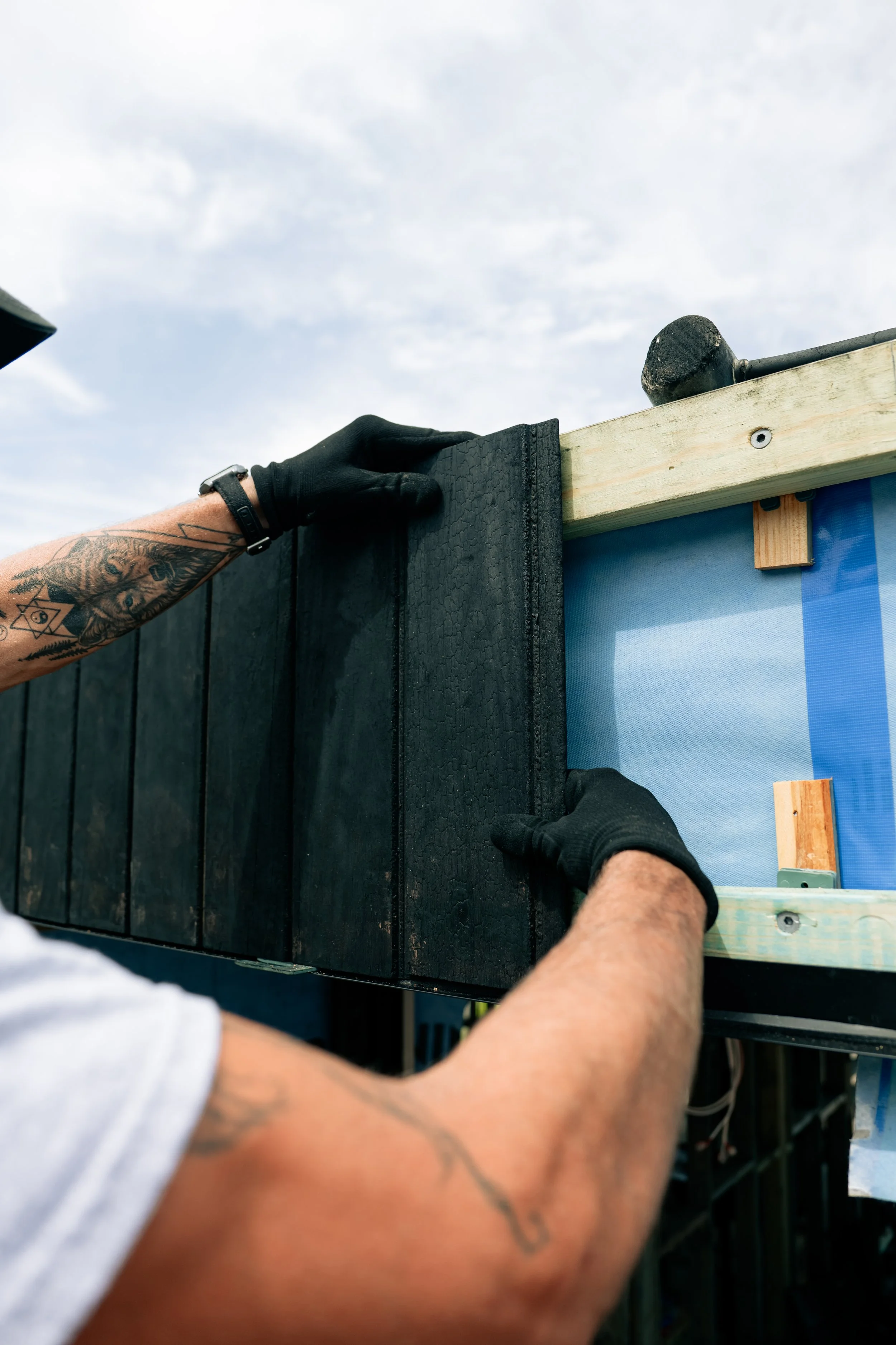 A person wearing black gloves is attaching a black wooden panel to a structure with real wood beams, outdoors under a partly cloudy sky.