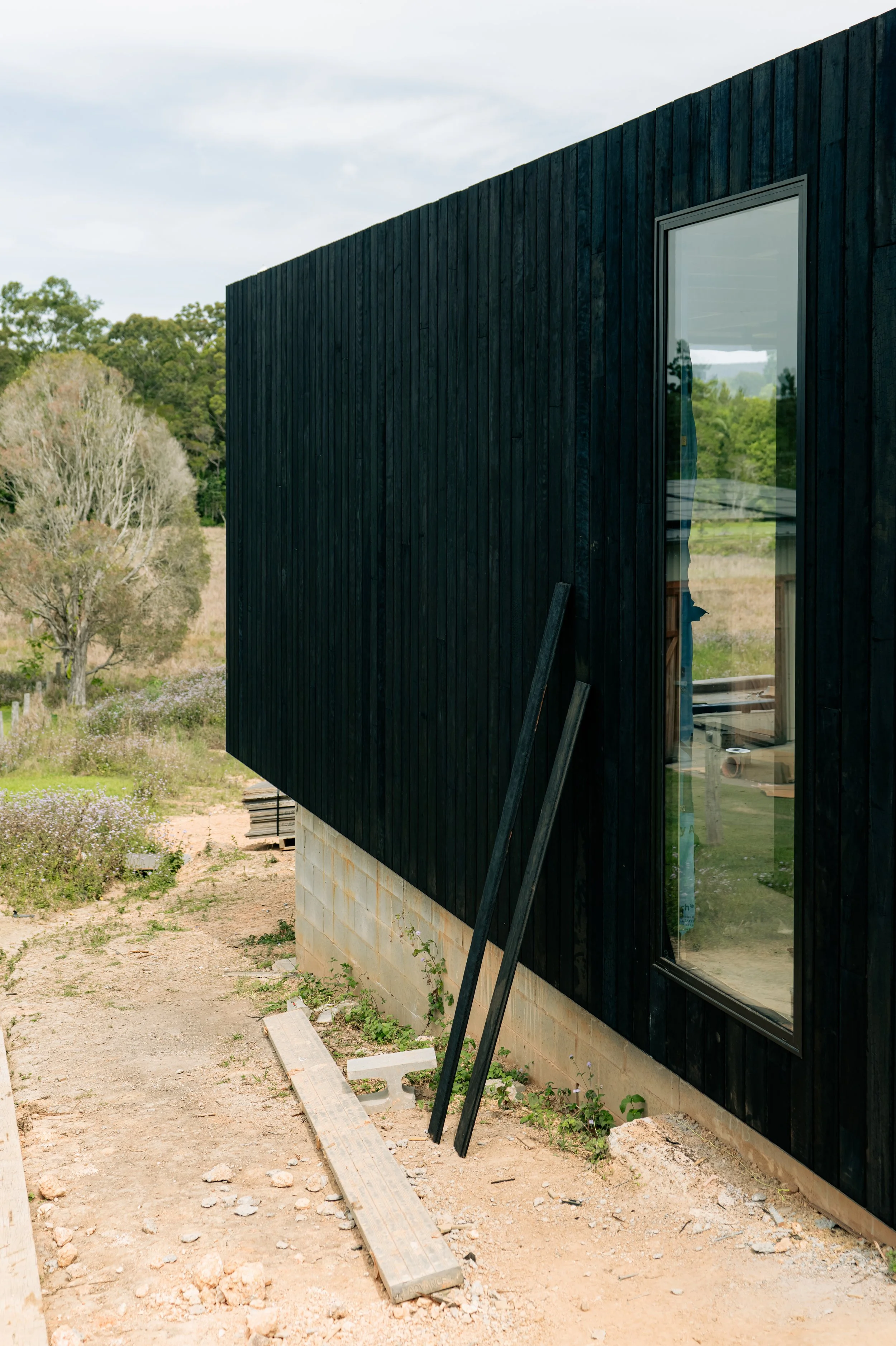 Part of a building with black wooden exterior cladding, a large vertical window, and construction materials on the ground outside in a rural area with trees in the background.