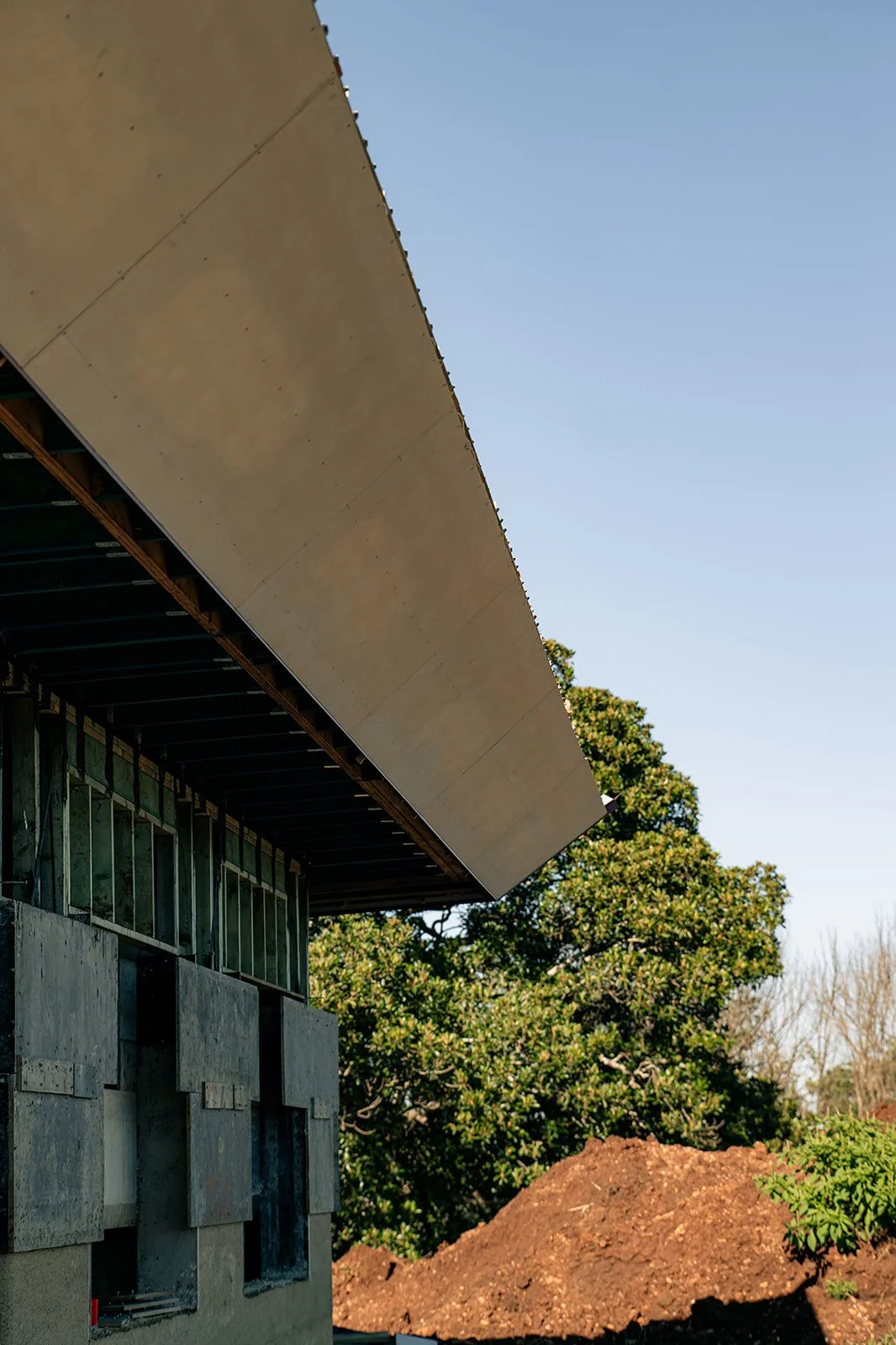 Partially constructed building with a sloped roof, concrete walls, and visible framing, set against a clear blue sky and trees.