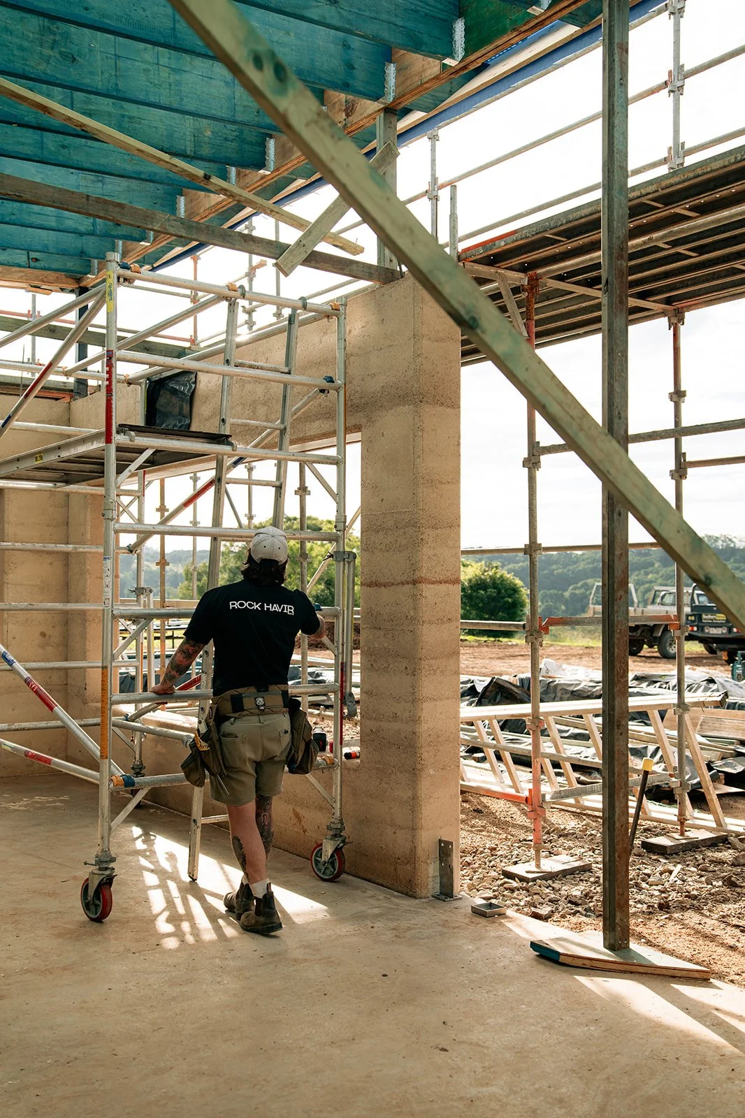 A construction worker walking through a partially built structure with scaffolding, with trees and construction equipment visible outside.