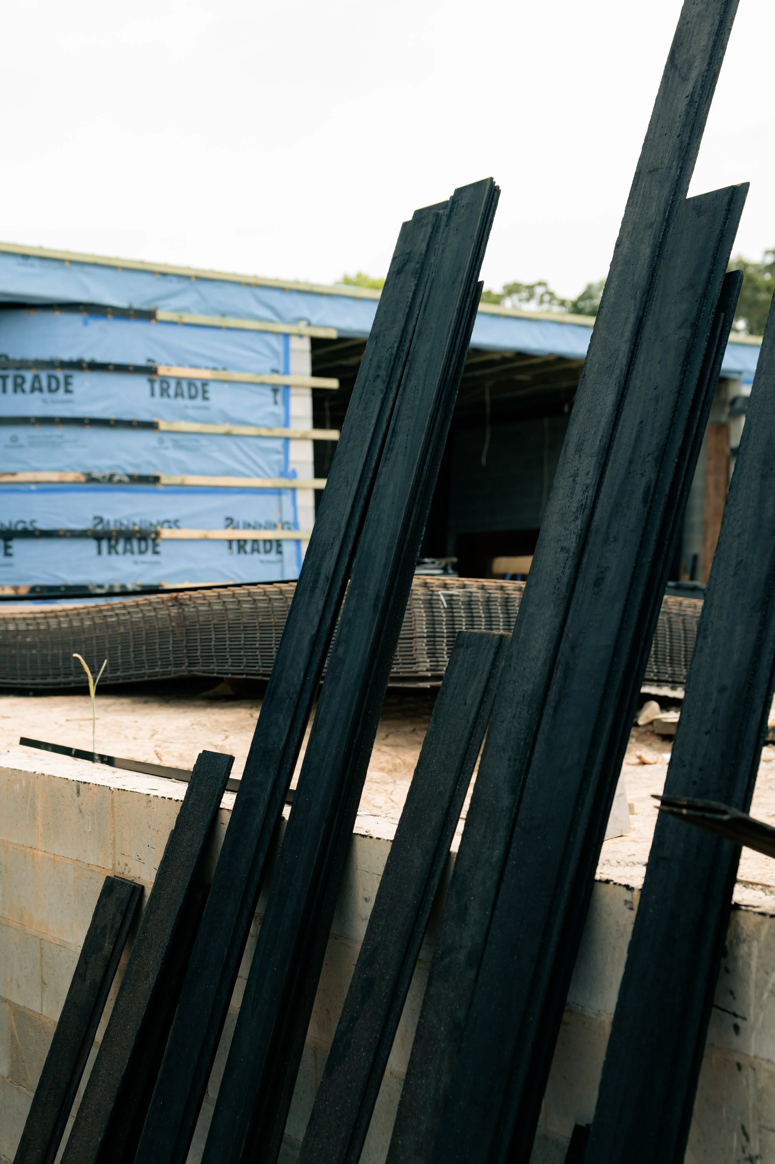 Close-up of black wooden planks leaning against a construction site wall with a house under construction in the background.
