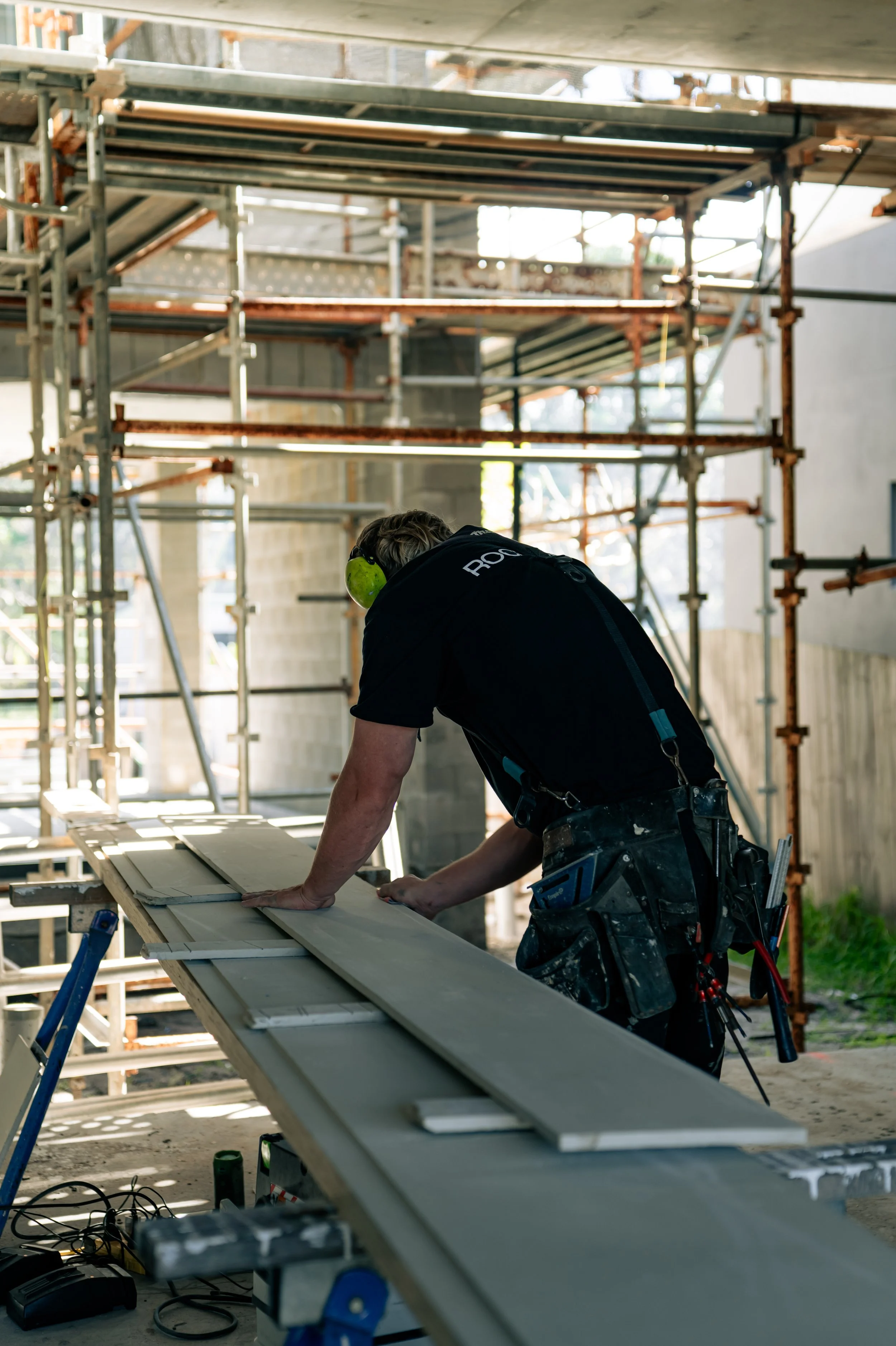 A construction worker in a black shirt and work belt with tools is working on a long wooden board at a construction site, surrounded by scaffolding.