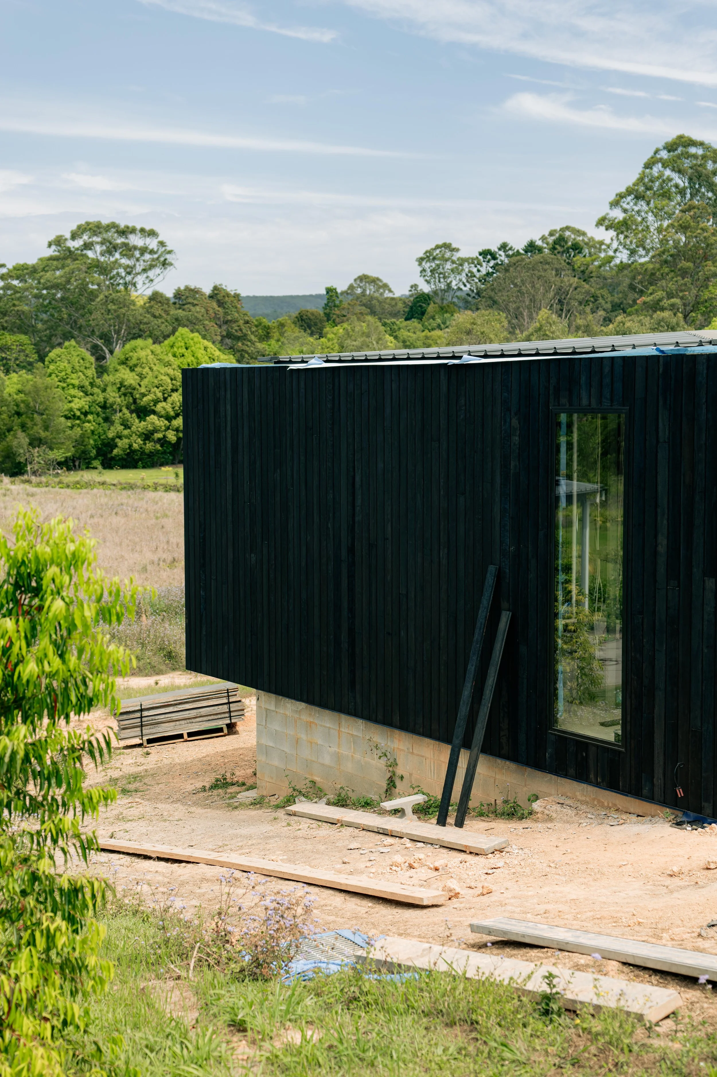Partial view of a black modern house under construction in a rural area, with construction materials and tools on ground, surrounded by trees and open landscape under a partly cloudy sky.