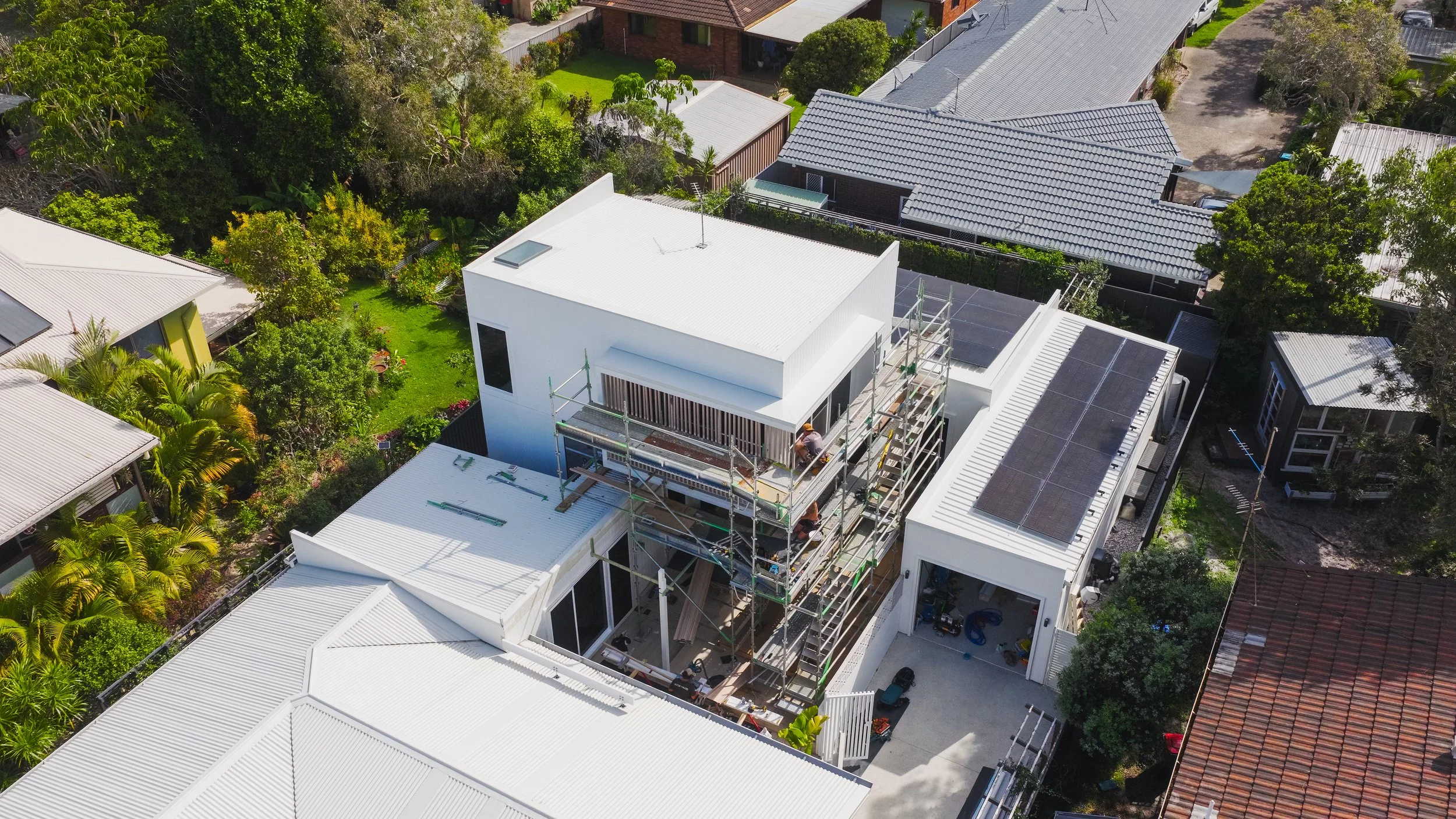 An aerial view of a modern white house under construction, with scaffolding around the upper levels. The house has solar panels on the roof and is surrounded by other homes with different roofing styles. Green trees and landscaped yards are visible around the neighborhood.