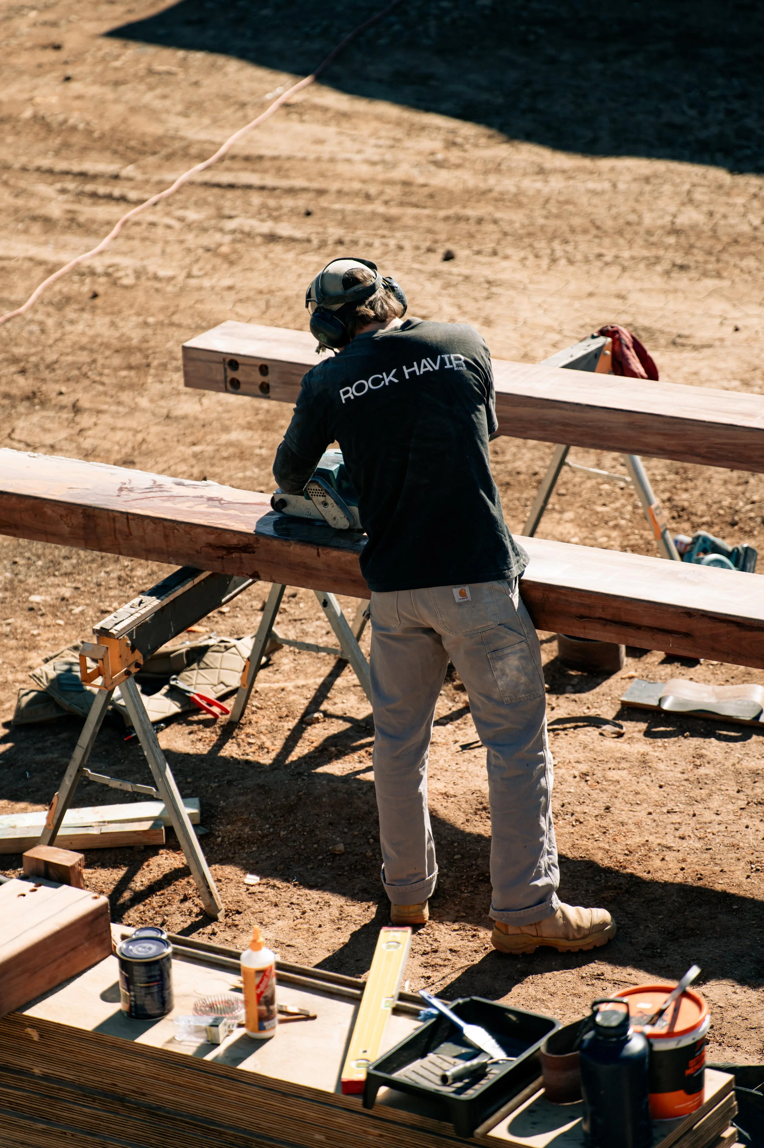 A person wearing a black shirt with 'ROCK HAVIR' written on the back, beige pants, and a helmet is sanding a large piece of wood outdoors, surrounded by tools and supplies.
