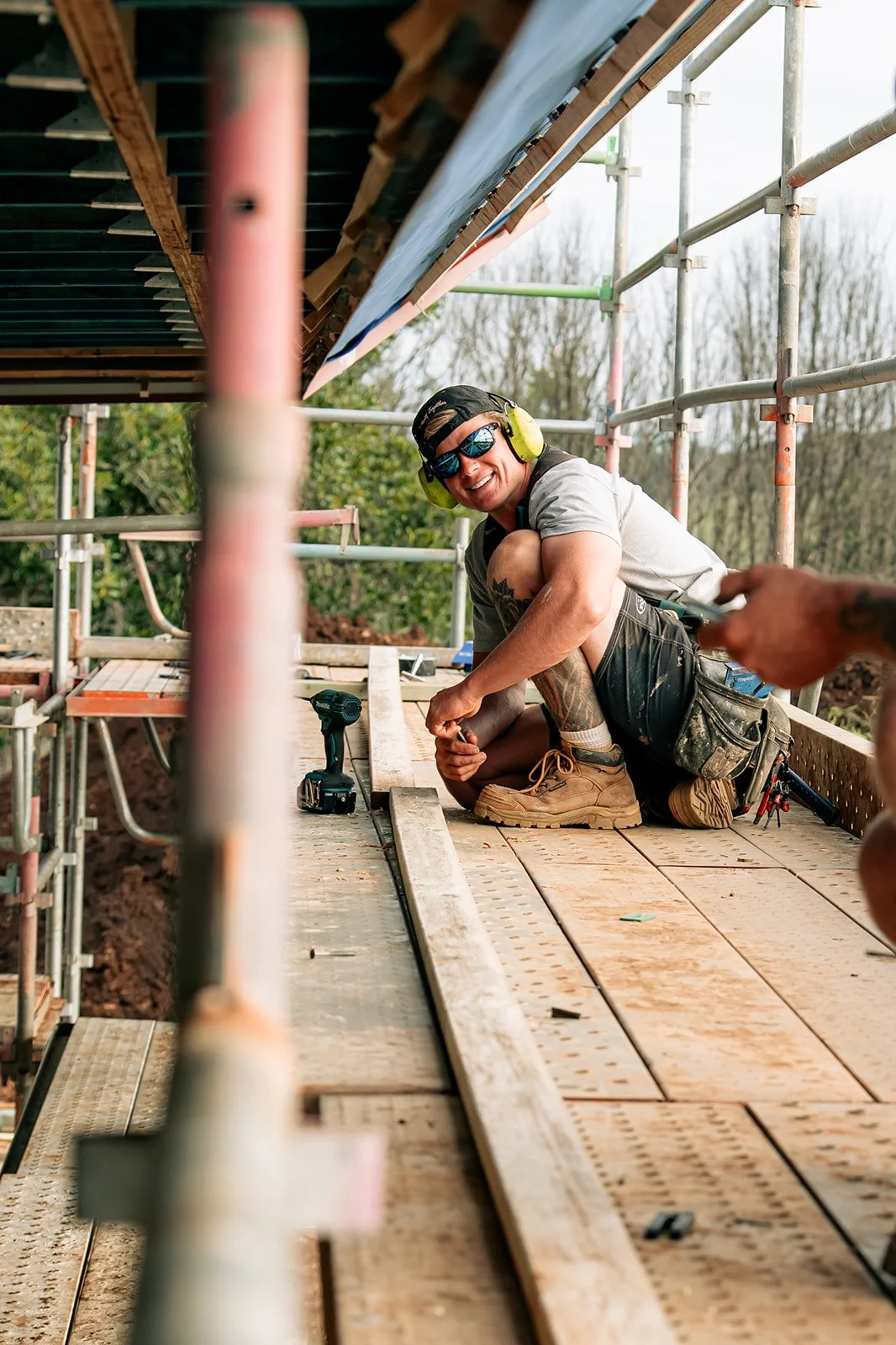 A smiling construction worker with sunglasses and hearing protection, kneeling on a scaffold while working on a wooden platform at an outdoor construction site.