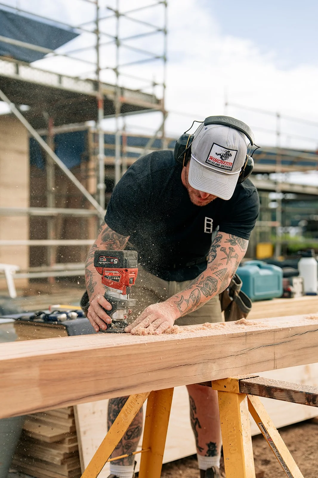 A man with tattoos working on woodworking at a construction site, using a power tool on a large wooden beam.