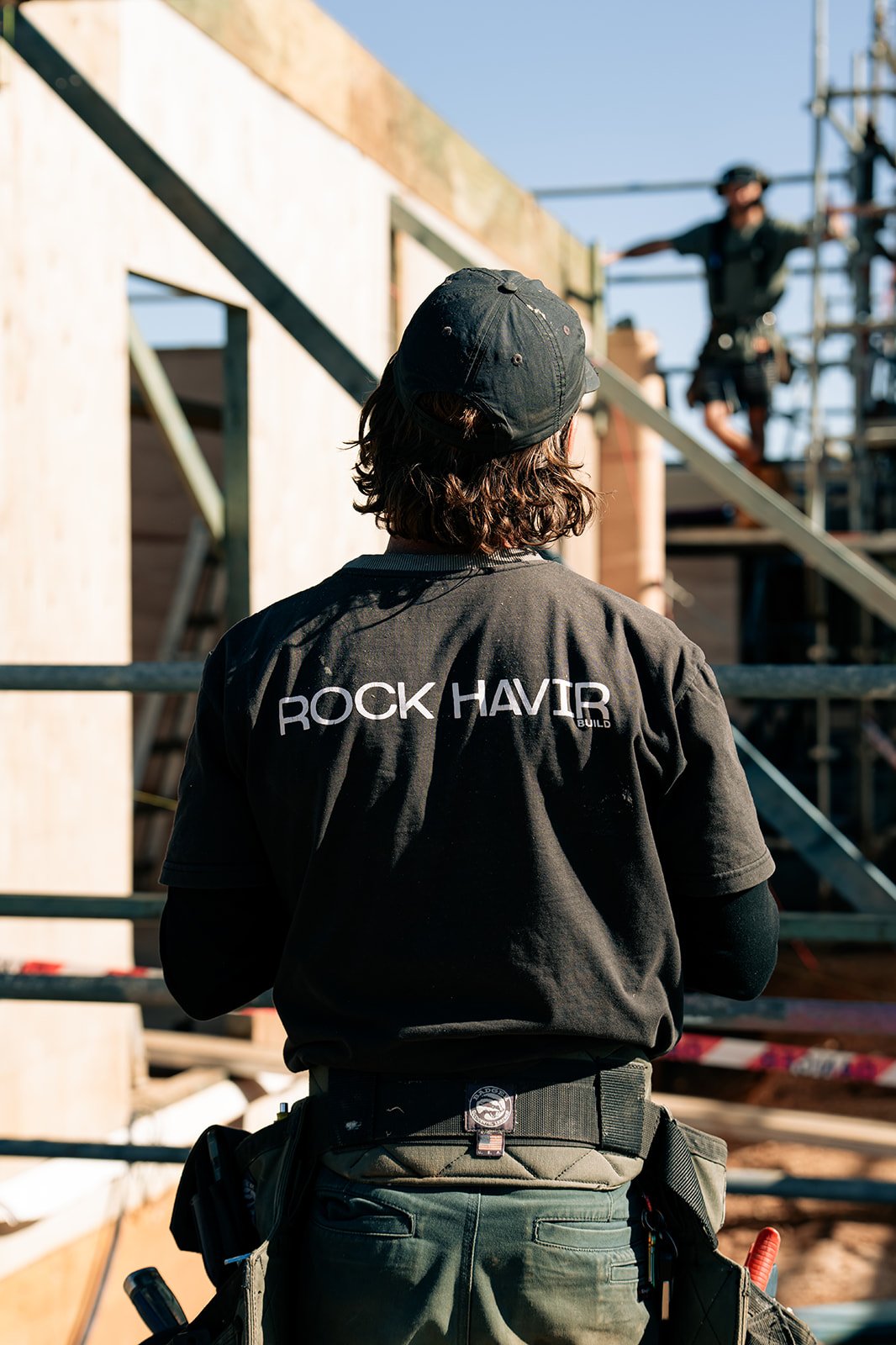 A construction worker with a back view, wearing a black shirt with 'ROCK HAVIR' written on it, working on a building site with scaffolding, and another worker is visible in the background.
