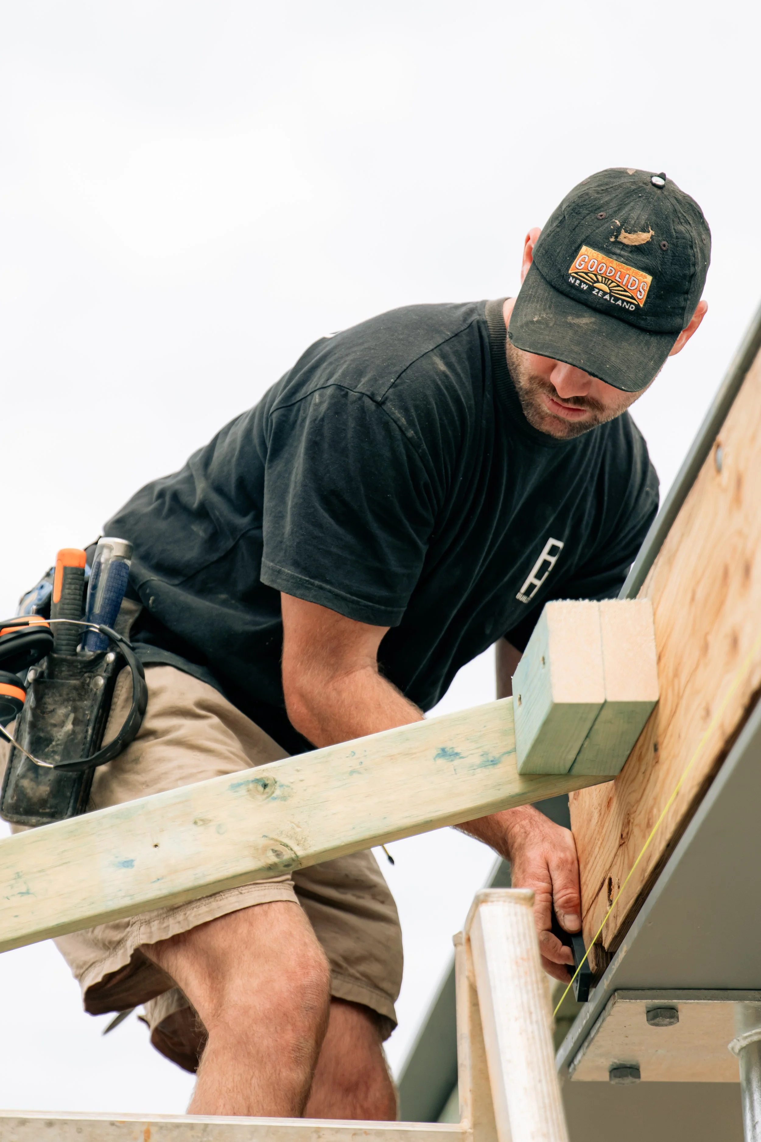 A man wearing a black cap, black t-shirt, and shorts is working on a construction project, securing a wooden beam to a wall.