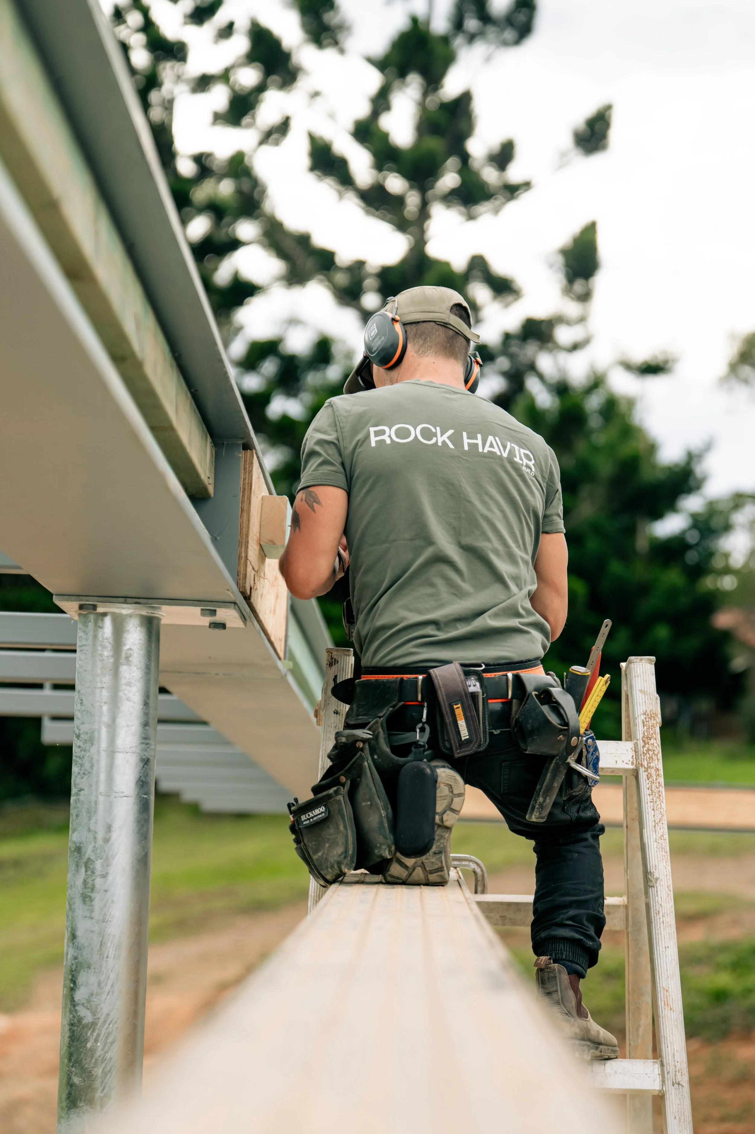 A construction worker wearing a green T-shirt with the words 'ROCK HAVIR' on the back, kneeling on a ladder, working on a construction project outdoors during daytime. The worker is wearing safety headphones and has tools attached to his belt.