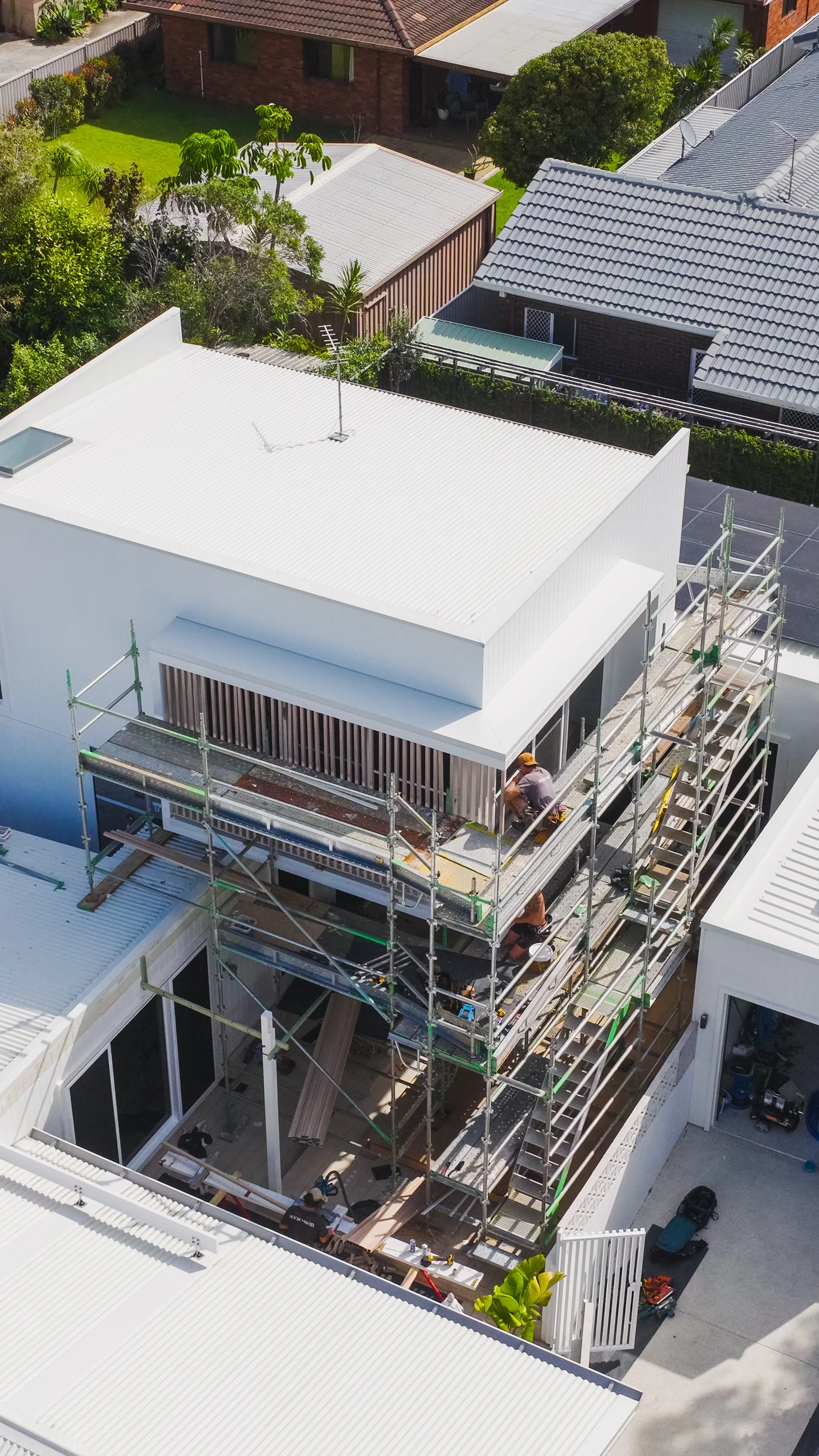 Construction workers working on scaffolding on a white modern house with a balcony, in a residential neighborhood with green trees and neighboring houses.