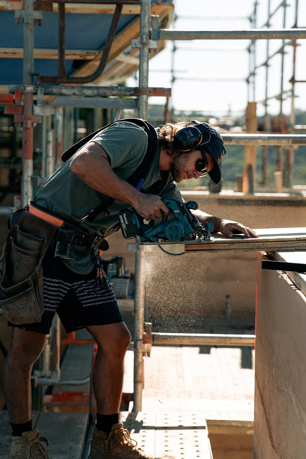 A construction worker using a circular saw to cut a wooden plank on a construction site with scaffolding and building materials around.