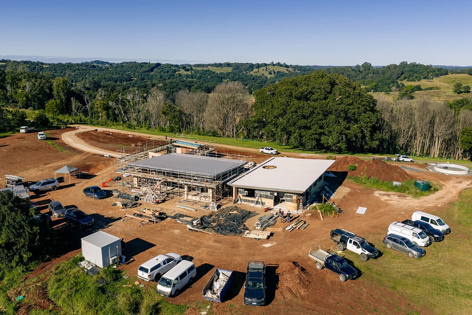 Aerial view of a construction site with buildings in progress, surrounded by trees and open land, with multiple parked vehicles nearby.