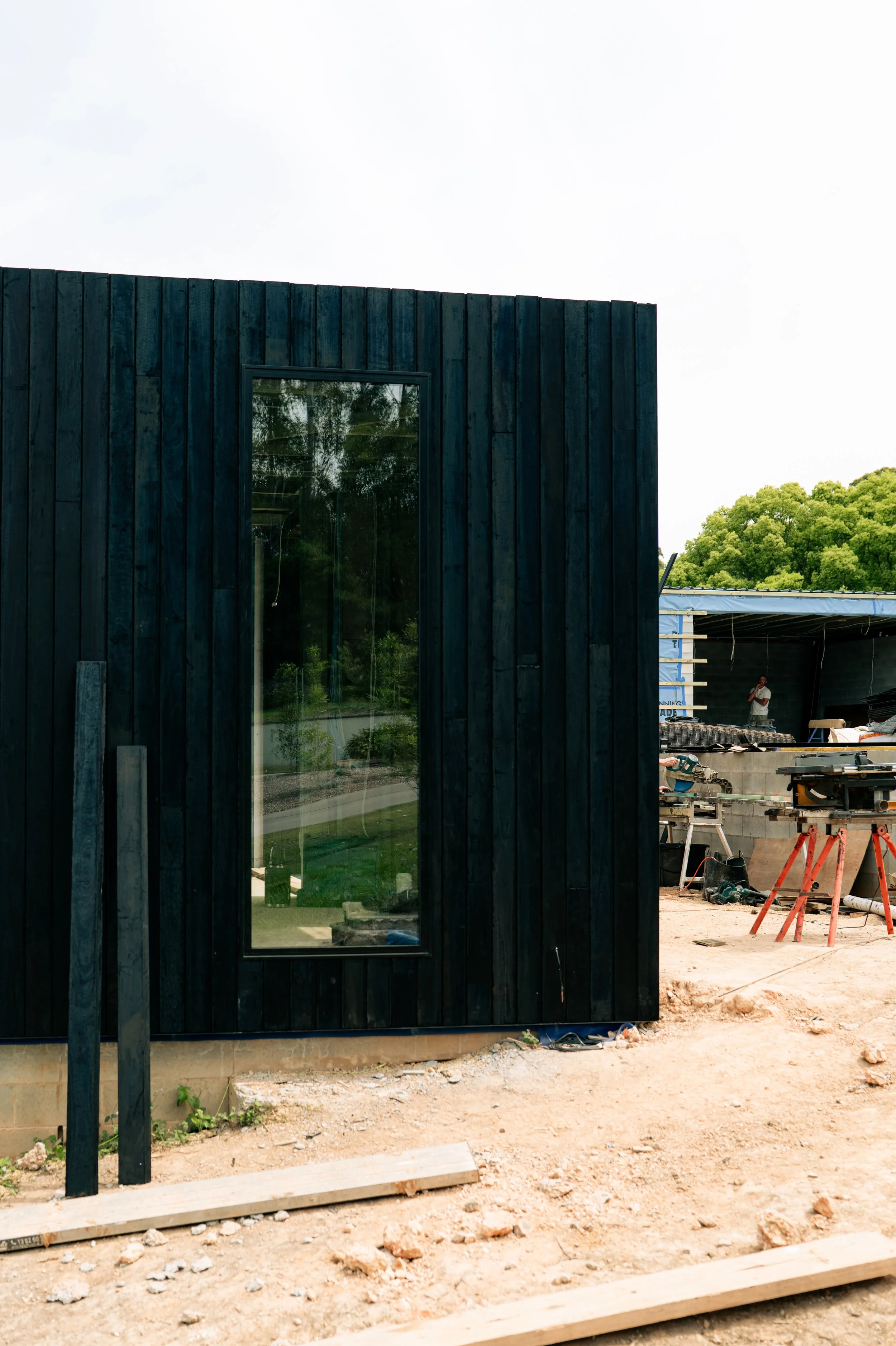 Part of a black wooden building under construction with a large glass window and construction tools nearby, in a setting with a green hill and a partly cloudy sky.