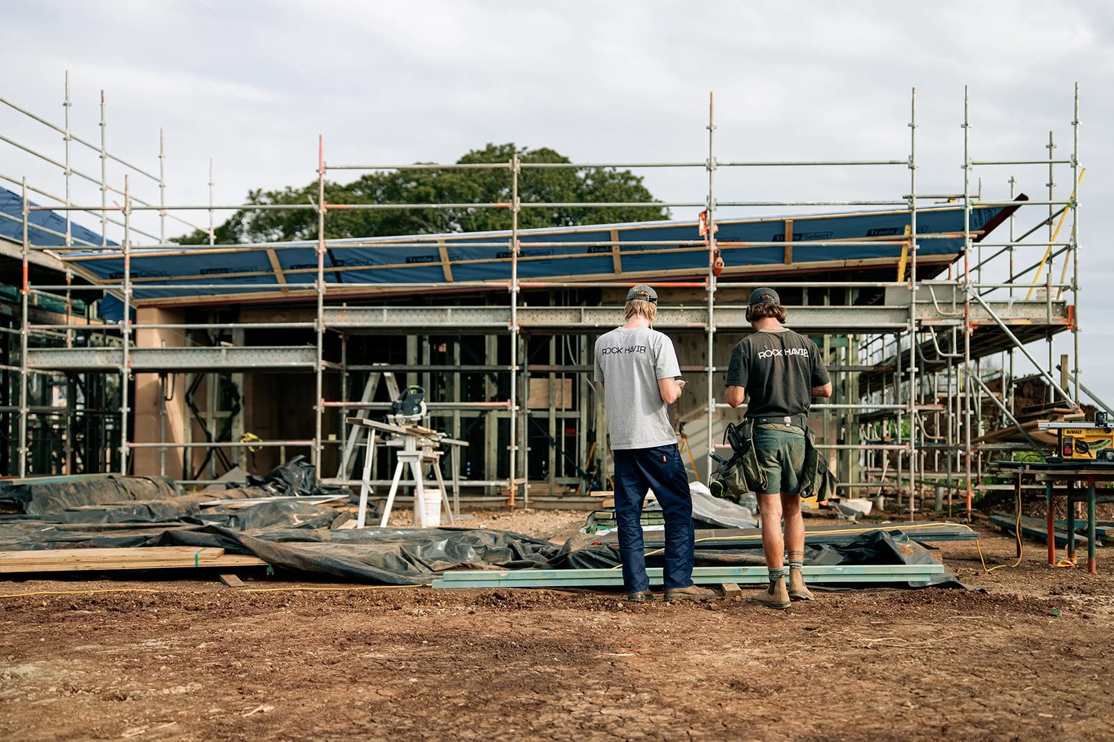 Two construction workers standing on a dirt foundation at a building site, with a partly constructed wooden structure and scaffolding behind them.