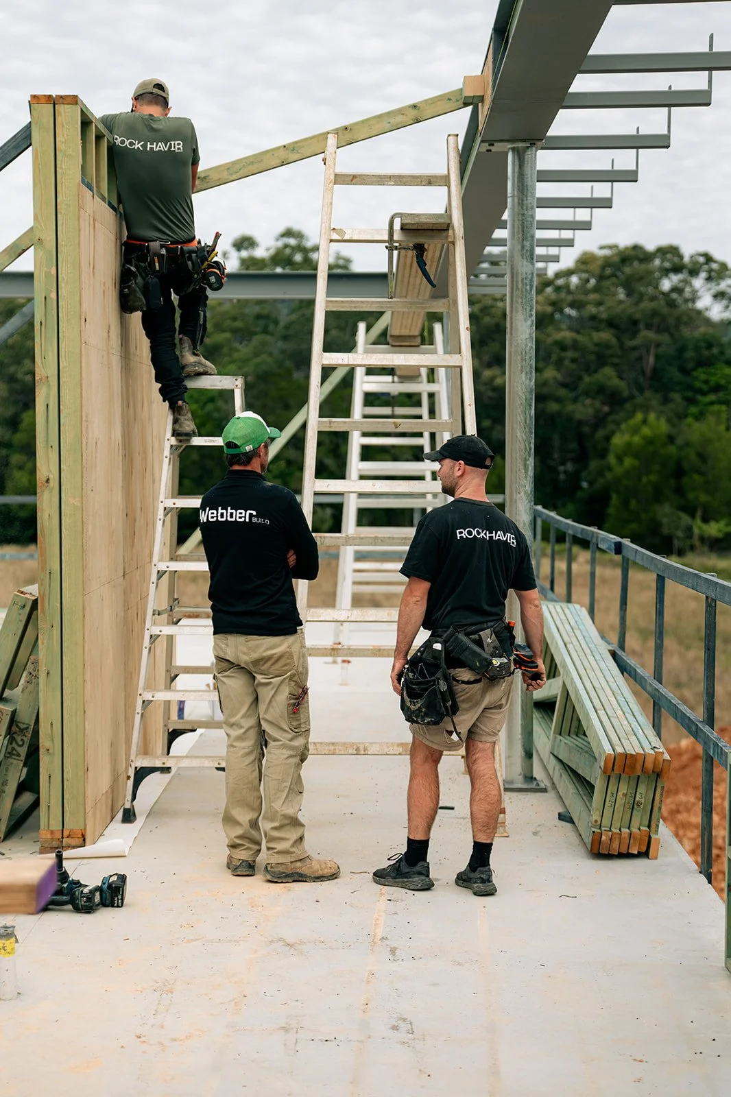 Three construction workers stand on a building site, with one working on framing and the other two observing and discussing. The structure is in the early stages with wooden and metal framing.
