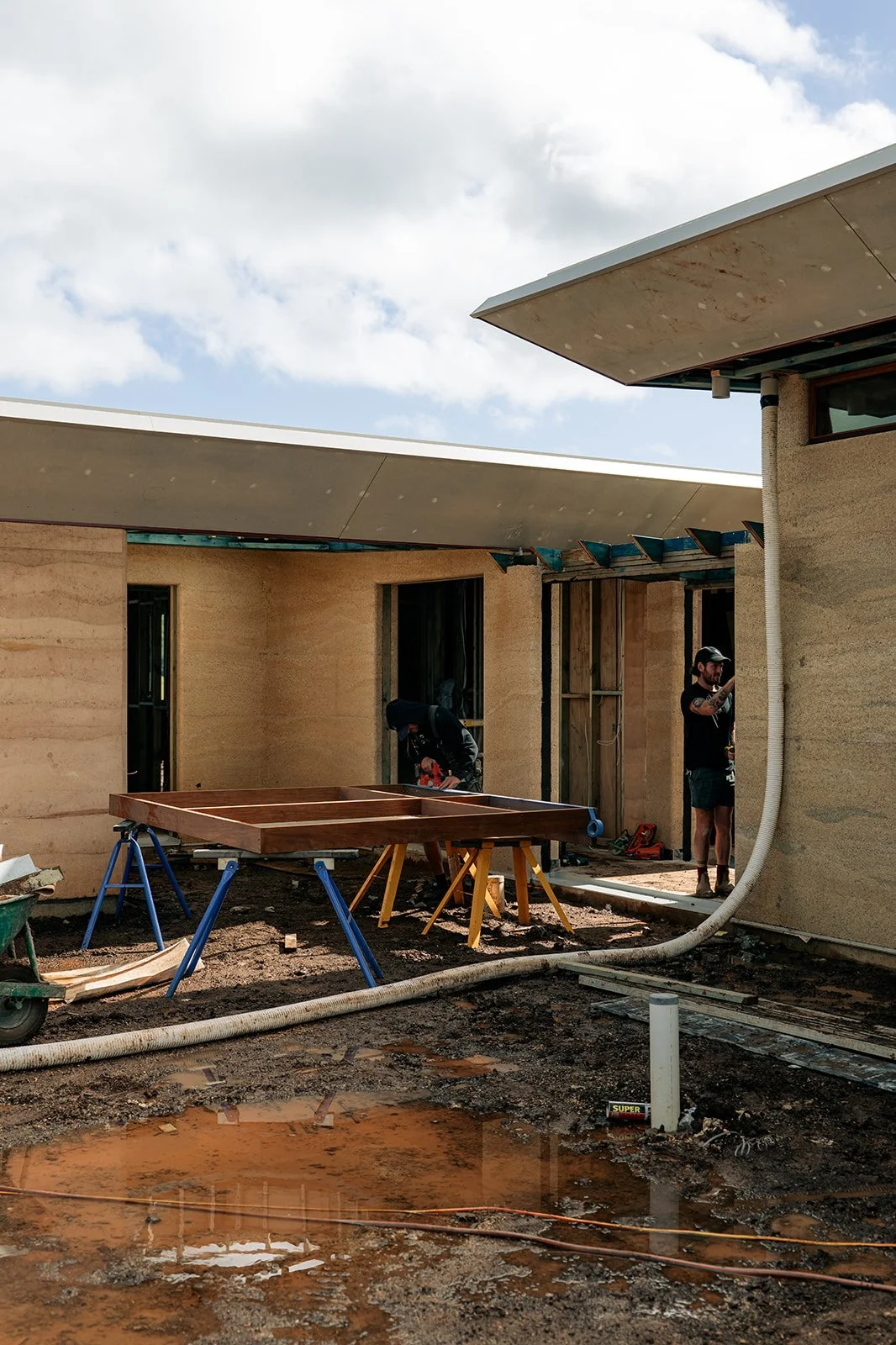 Construction site with workers installing a wooden structure outside a house. The scene includes mud, construction tools, and partially built walls with a cloudy sky overhead.