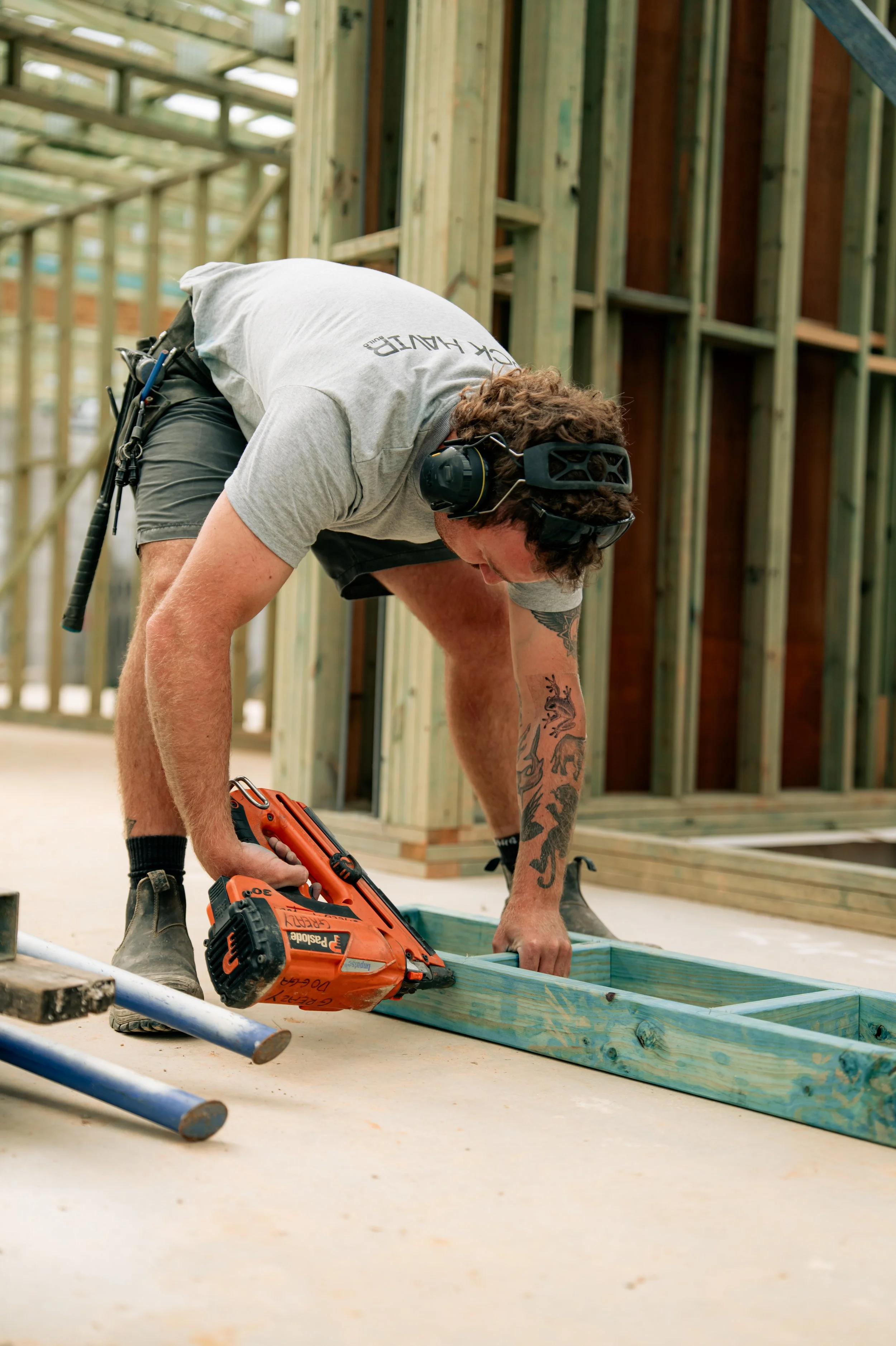 Man working on a construction site, cutting a piece of wood with a nail gun. He is wearing hearing protection and is surrounded by wooden framing structures.