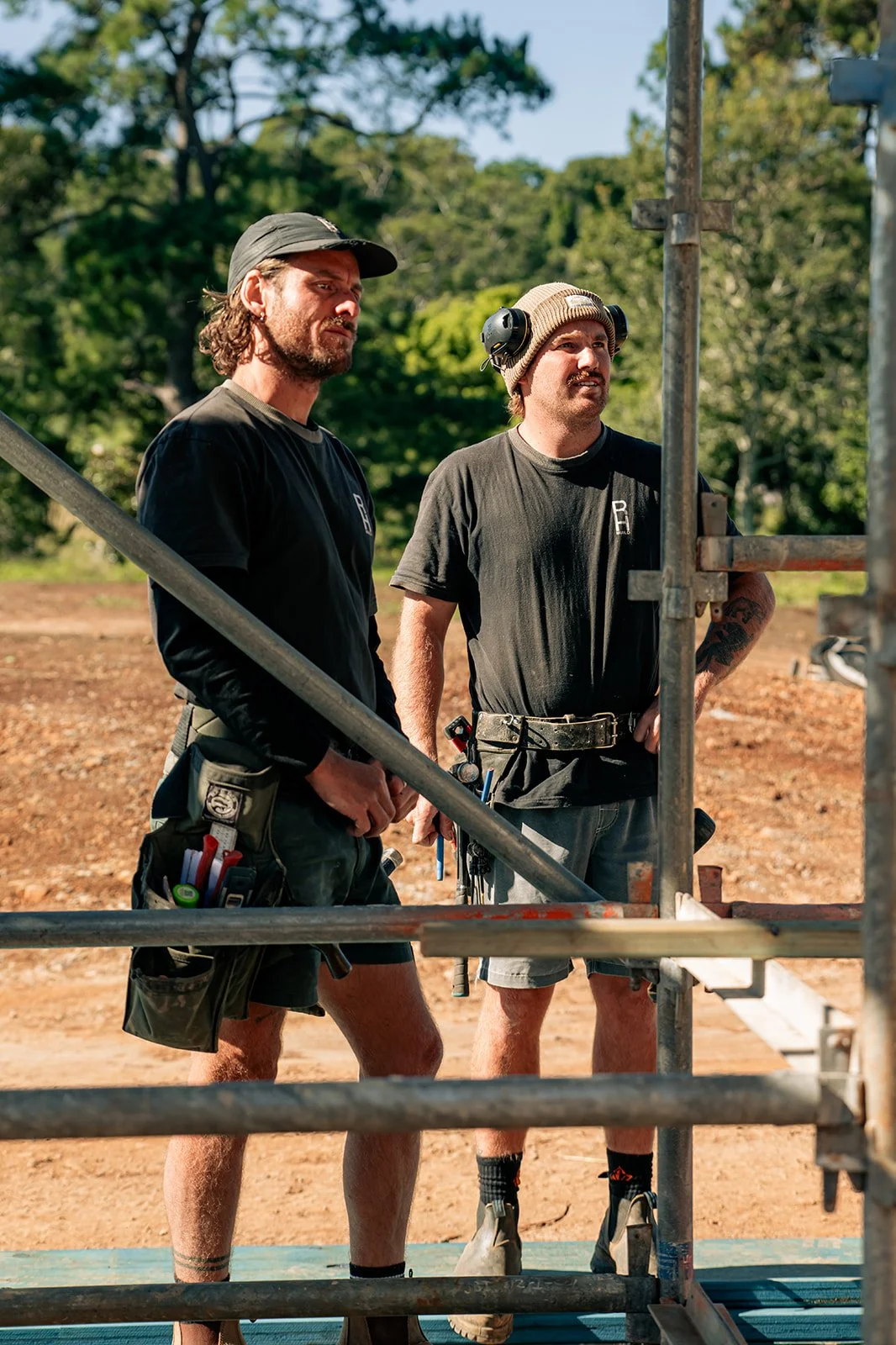 Two construction workers standing on a construction site, looking at scaffolding. One has long hair and is wearing a black cap, the other has a beanie and is wearing hearing protection around his neck.