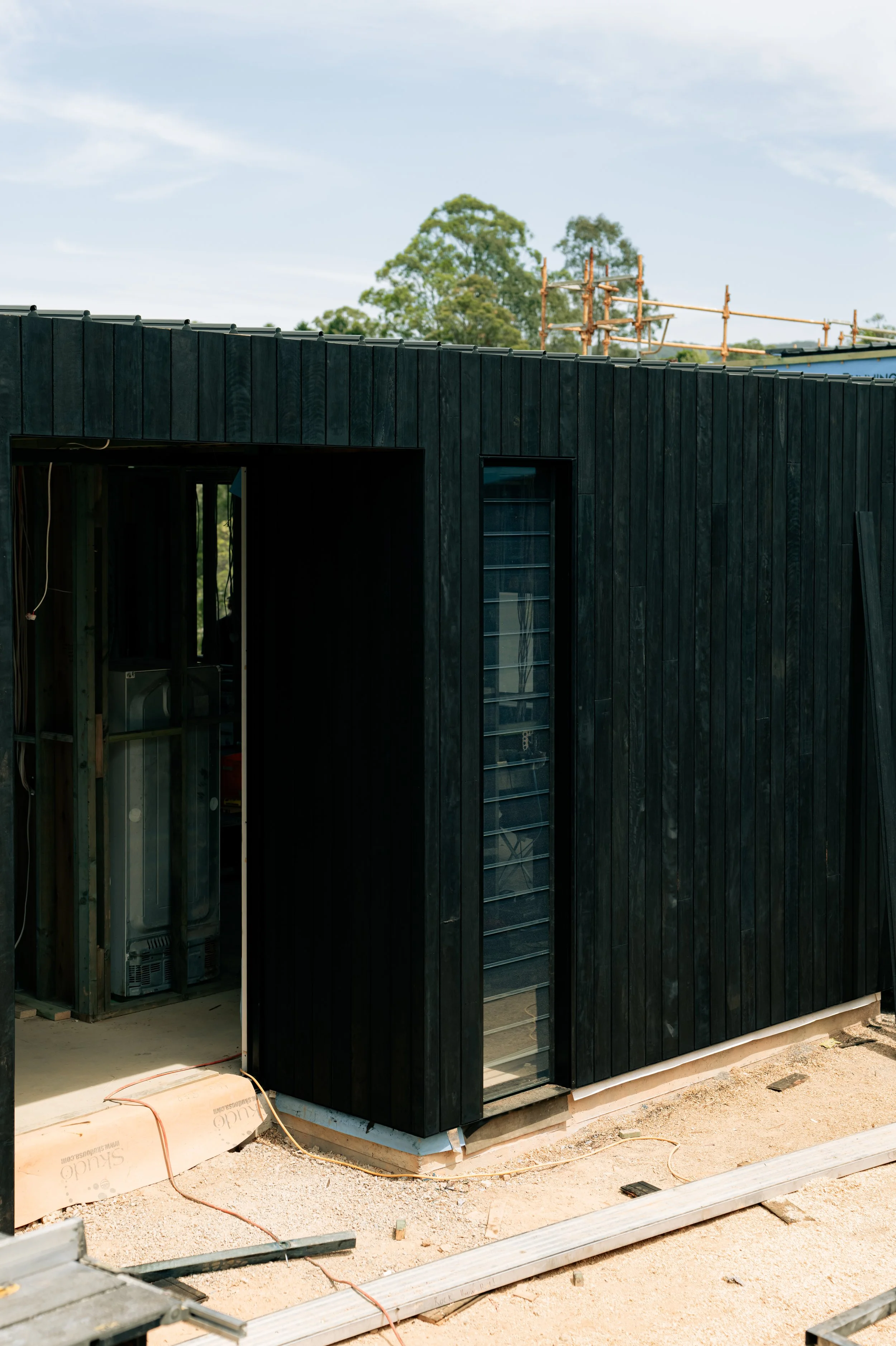 Exterior of a building under construction with black cladding, a partially open doorway, and a window. Construction materials and tools are on the ground.