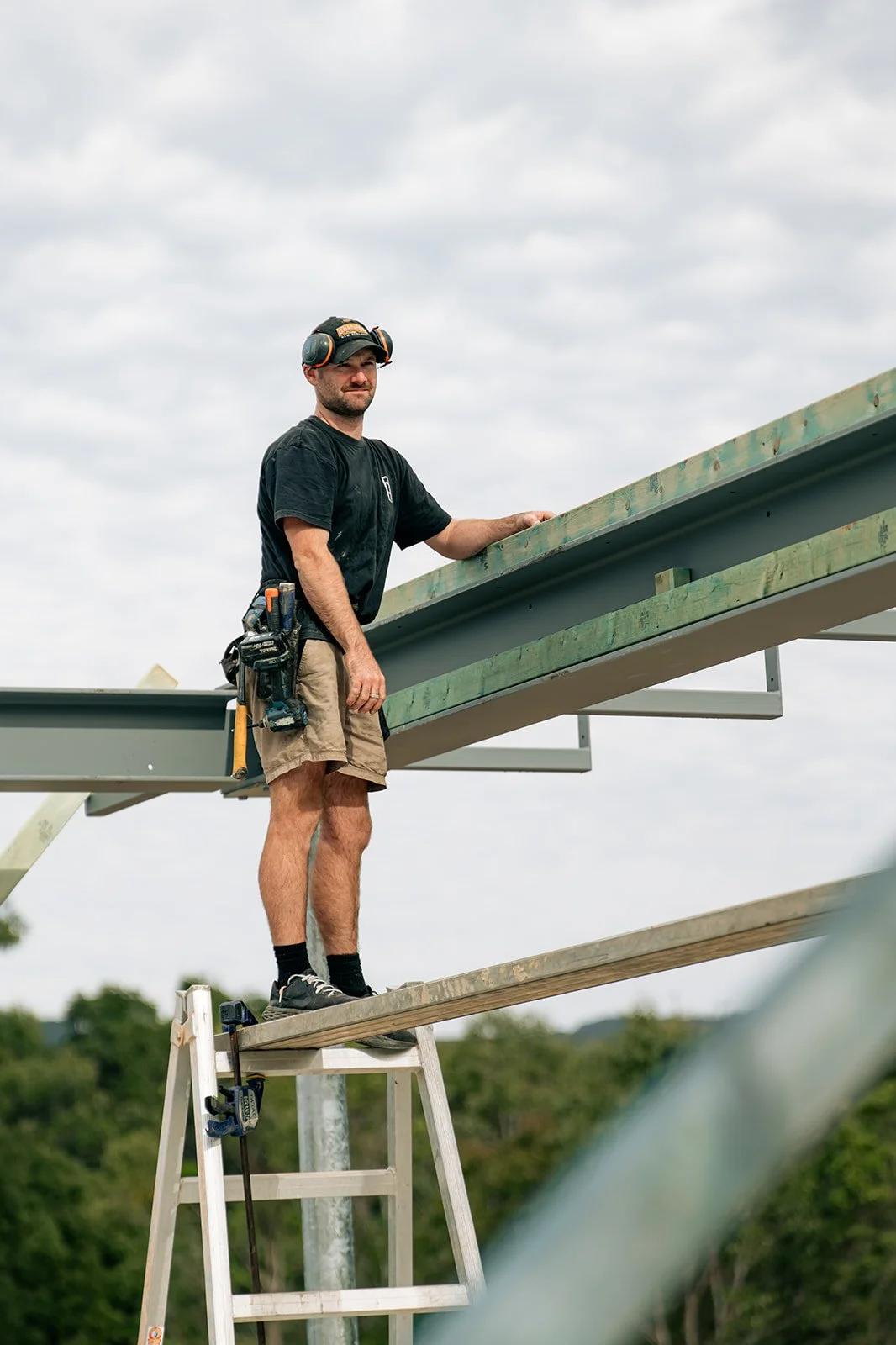 A man wearing a black t-shirt, tan shorts, and safety gear, standing on a ladder, working on a construction project with metal beams against a cloudy sky background.