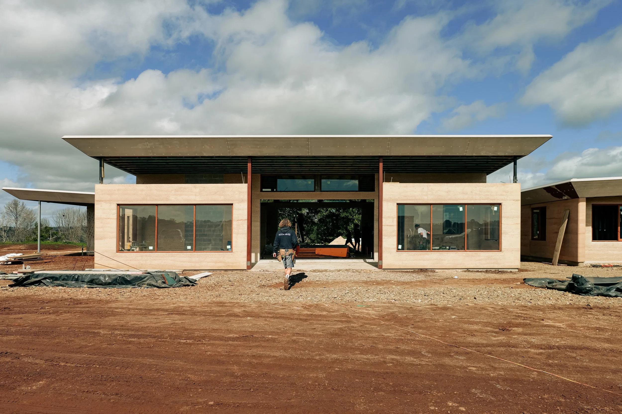 Modern house under construction with a flat roof and large windows, construction workers present, dirt ground, cloudy sky.