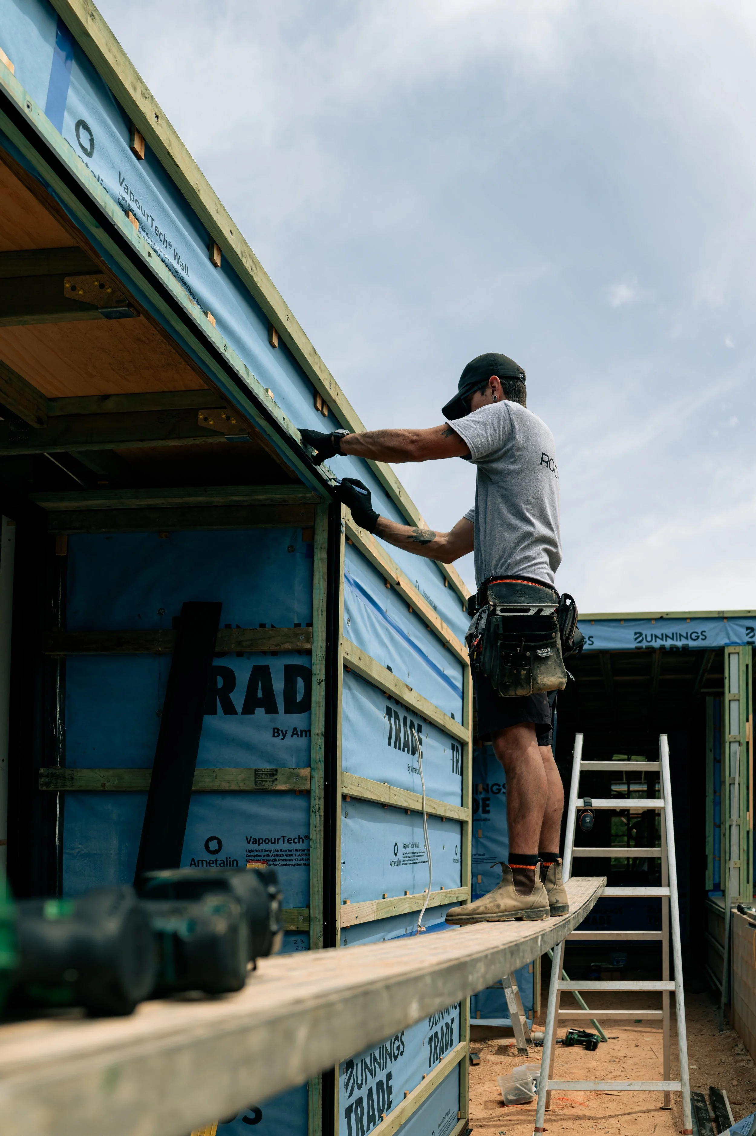 A construction worker standing on a ladder, installing the exterior cladding of a building under construction, with a cloudy sky in the background.