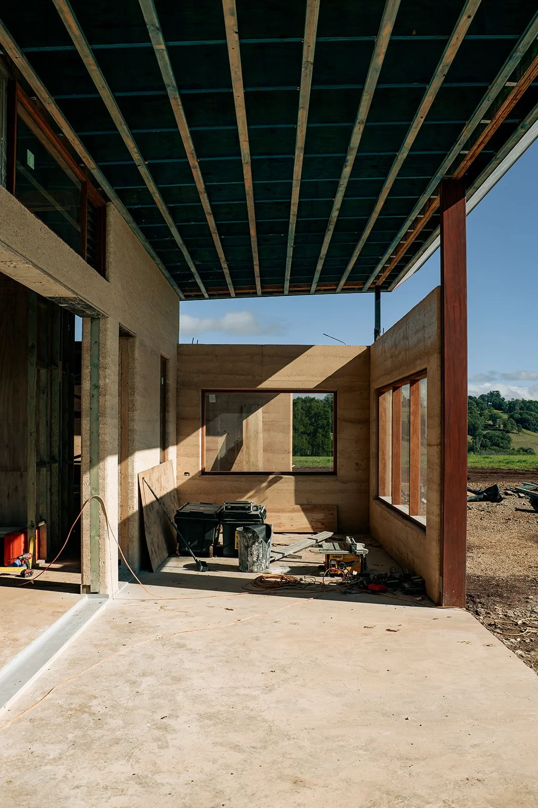Partial view of a building under construction with wooden framework, large windows, and construction tools on the floor, overlooking a green landscape.