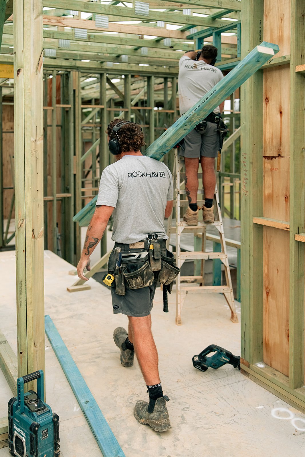 Two construction workers assembling a wooden structure, with one worker walking and the other on a ladder, carrying a long wooden beam.