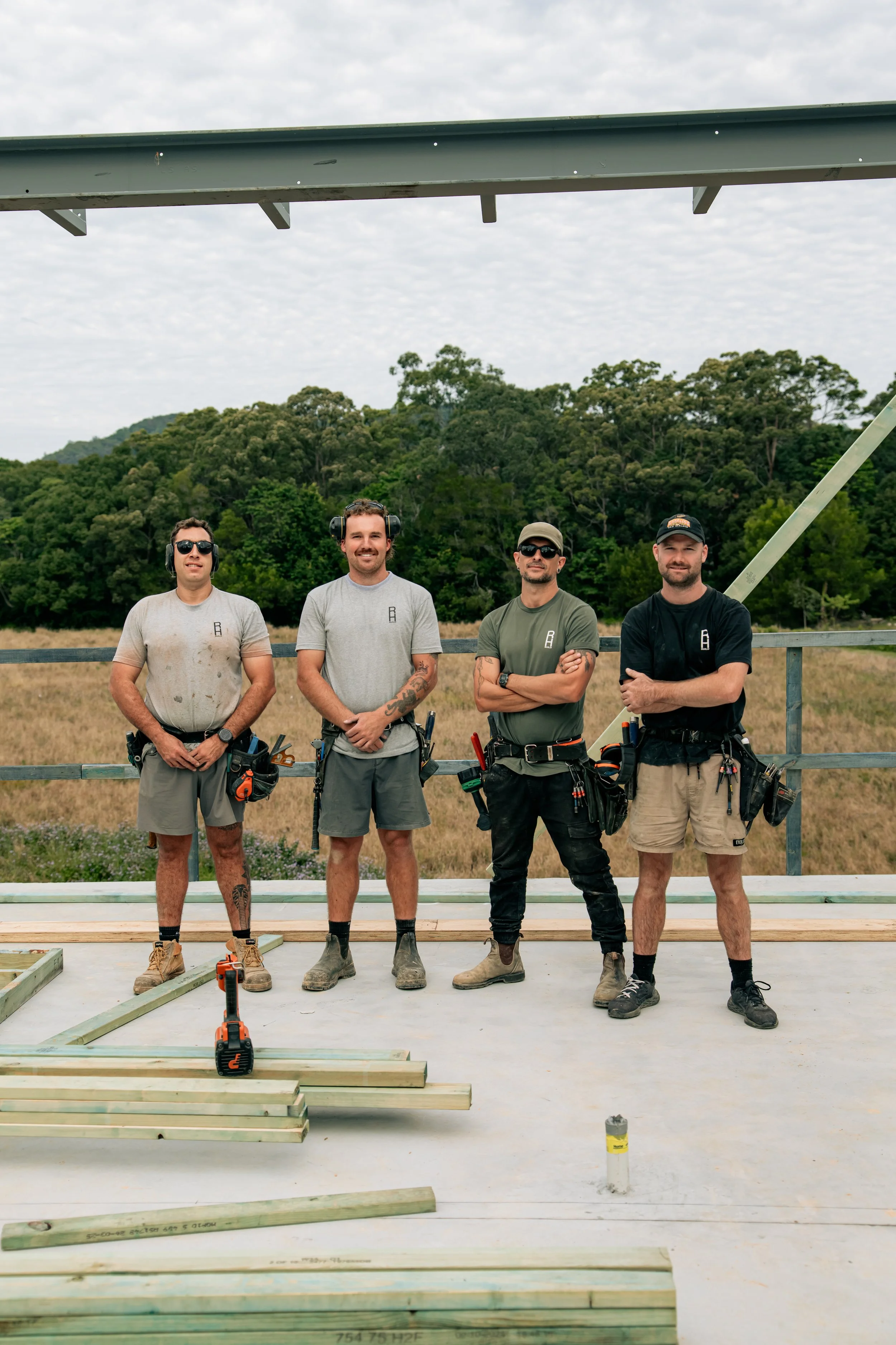 Four construction workers standing on a concrete floor with construction tools and wooden beams, outdoors with a backdrop of trees and cloudy sky.