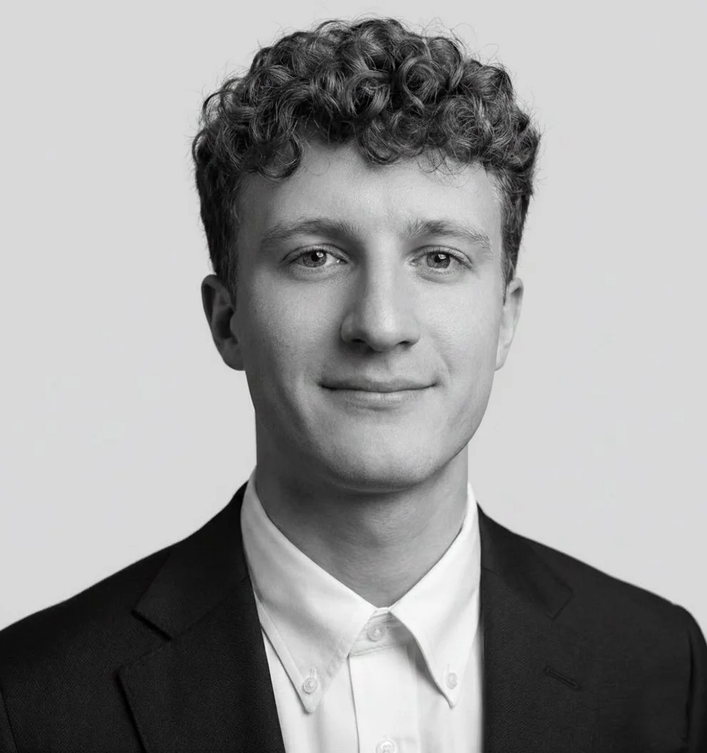 Black and white headshot of a young man with curly hair wearing a suit and white shirt.
