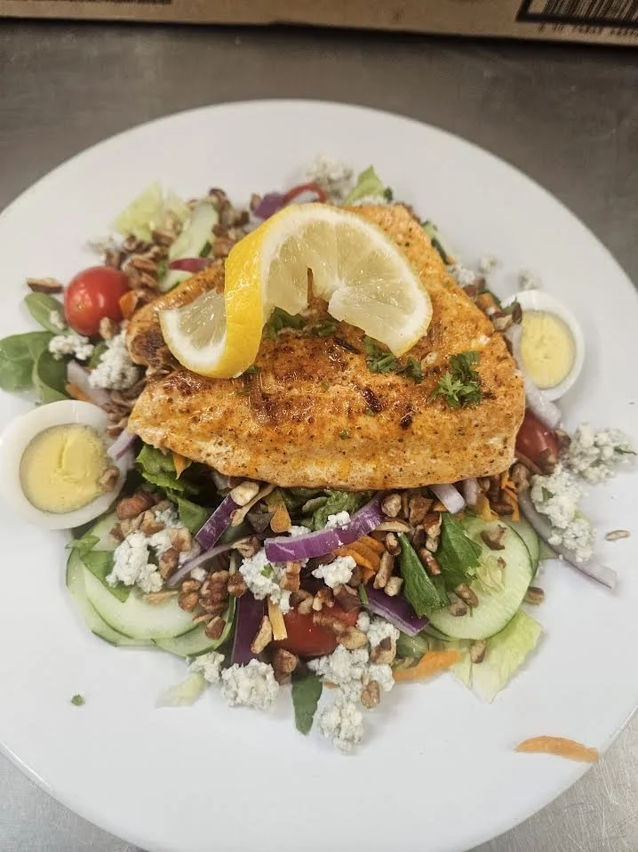 Salad with grilled salmon, lemon slices, boiled eggs, cherry tomatoes, cucumbers, red onions, shredded carrots, blue cheese crumbles, chopped nuts, and fresh herbs.