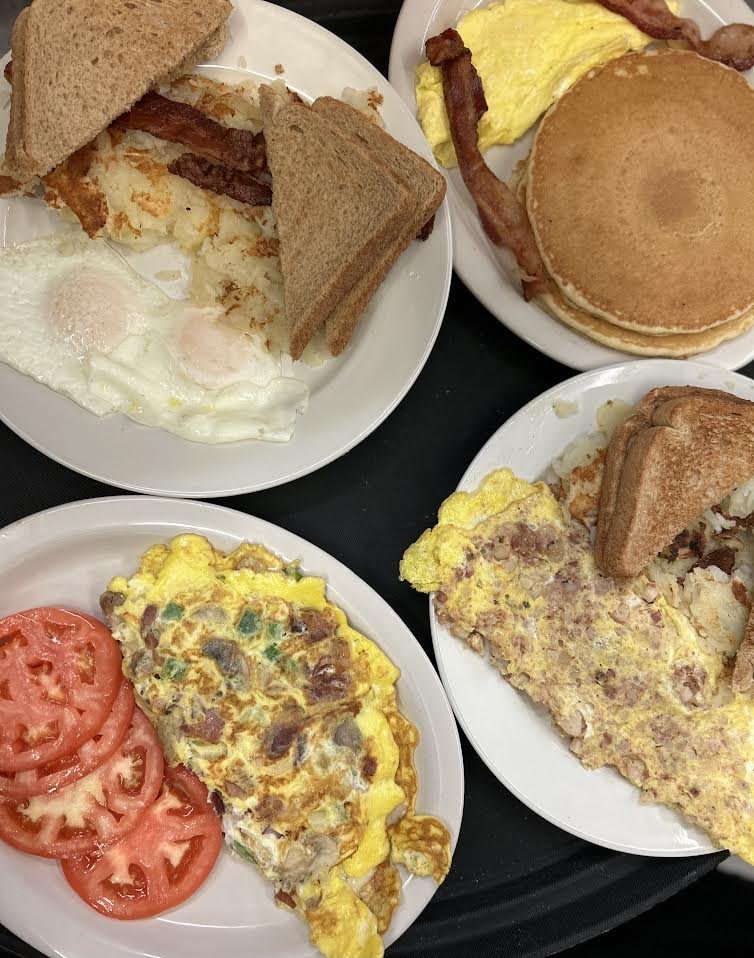 A breakfast spread with plates of scrambled eggs with vegetables, sliced tomatoes, pancakes with bacon, a breakfast sandwich, and an omelette.