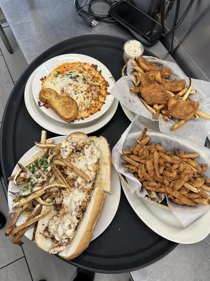 Tray with a slice of pizza with cheese and herbs, a toasted garlic bread, a fish sandwich with French fries and green herbs, fried chicken with French fries, and a basket of curly fries, on a black tray with a small container of dipping sauce.