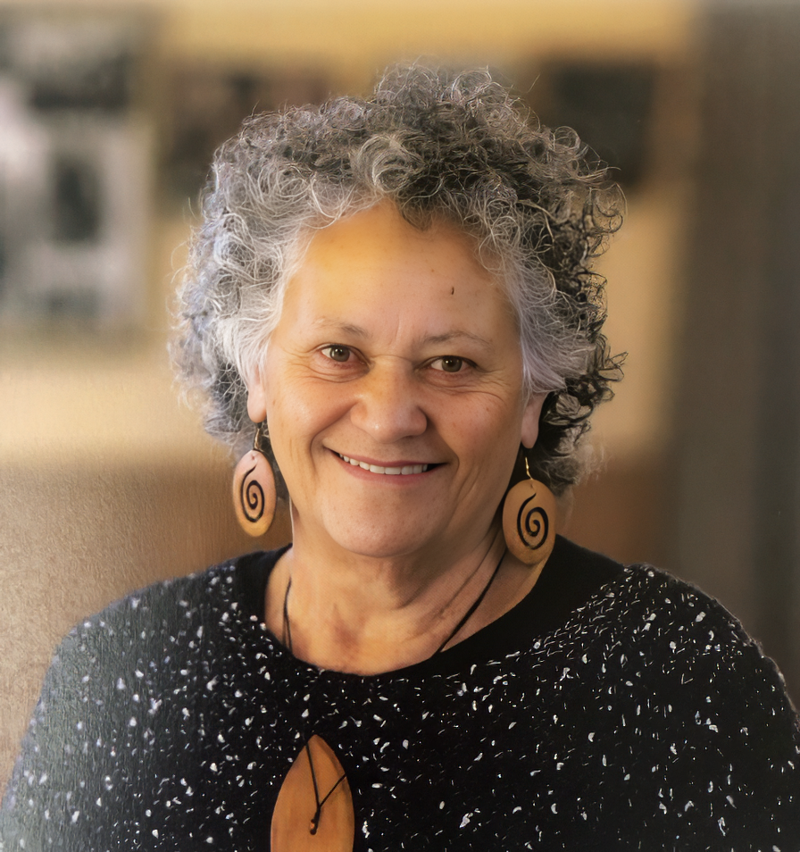 A smiling woman with curly gray and black hair, wearing wooden spiral earrings and a black sweater with white specks, sitting indoors.