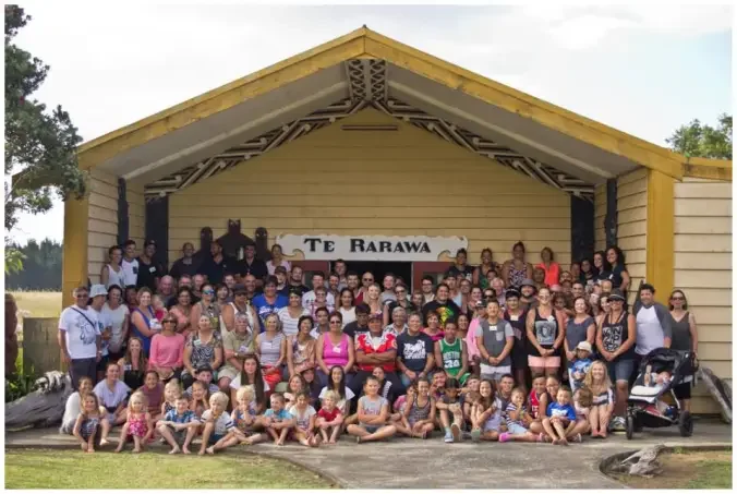 Group of people, including children, gathered on a stage with a sign that reads 'Te Rarawa,' in front of a wooden building with a triangular roof.