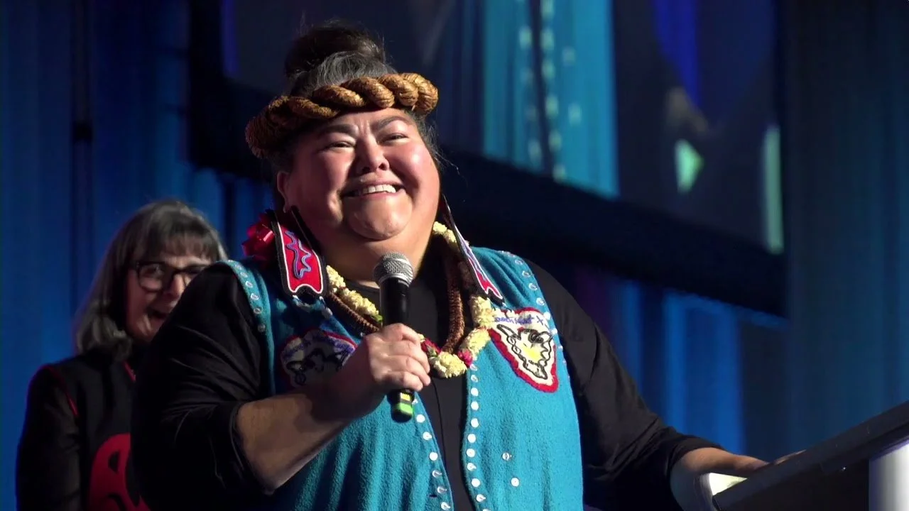 A woman wearing traditional attire, with braided hair on her head, holding a microphone and smiling on stage. Two women are in the background.