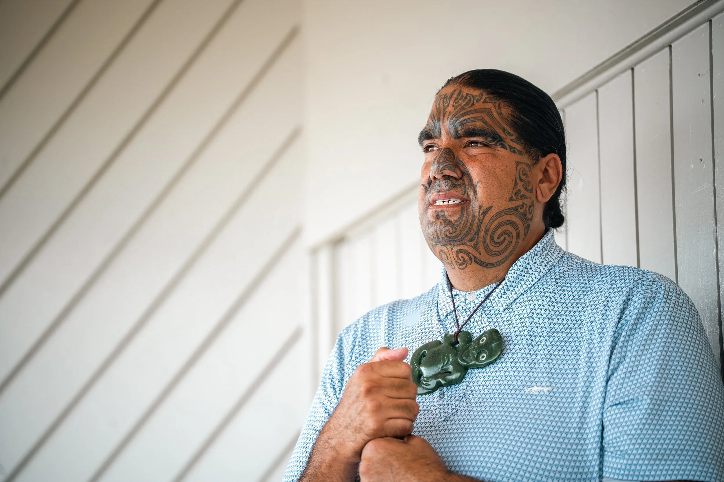 A man with traditional Maori facial tattoos, wearing a black shirt with a logo, sitting indoors with a plant and abstract art in the background.