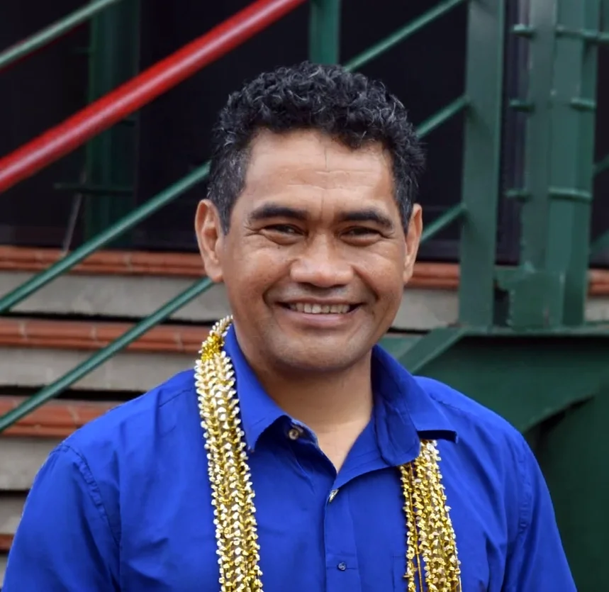 A man smiling, wearing a blue shirt and gold garlands, standing outdoors in front of a staircase with green and red railing.
