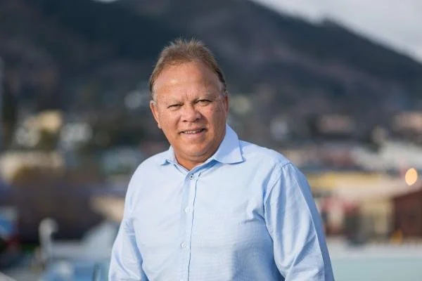 A man with light skin and blonde hair standing outdoors in front of a blurred cityscape and mountains, wearing a light blue button-up shirt.
