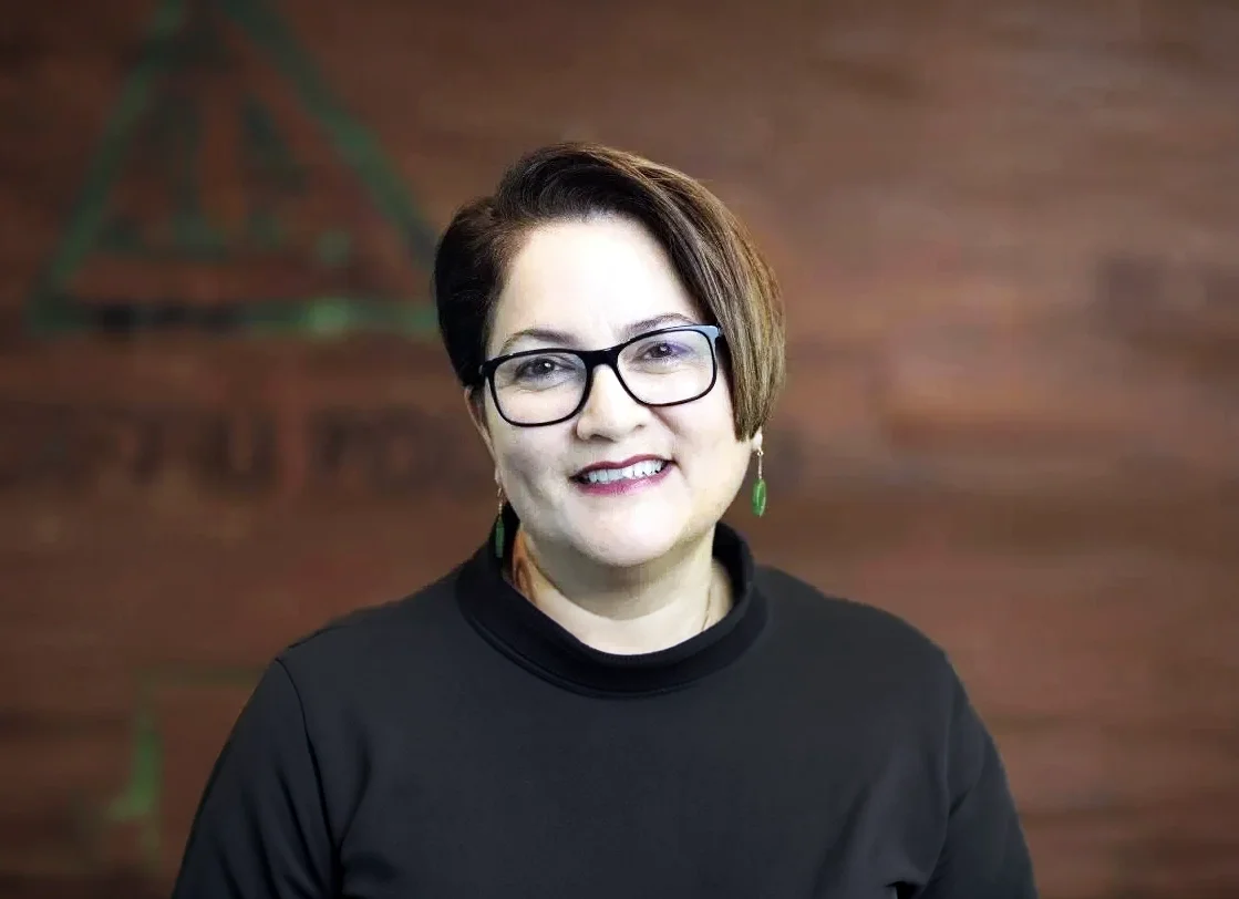 A woman with short brown hair, glasses, and green earrings, smiling and wearing a black top, standing in front of a wooden wall with a faint logo or design.