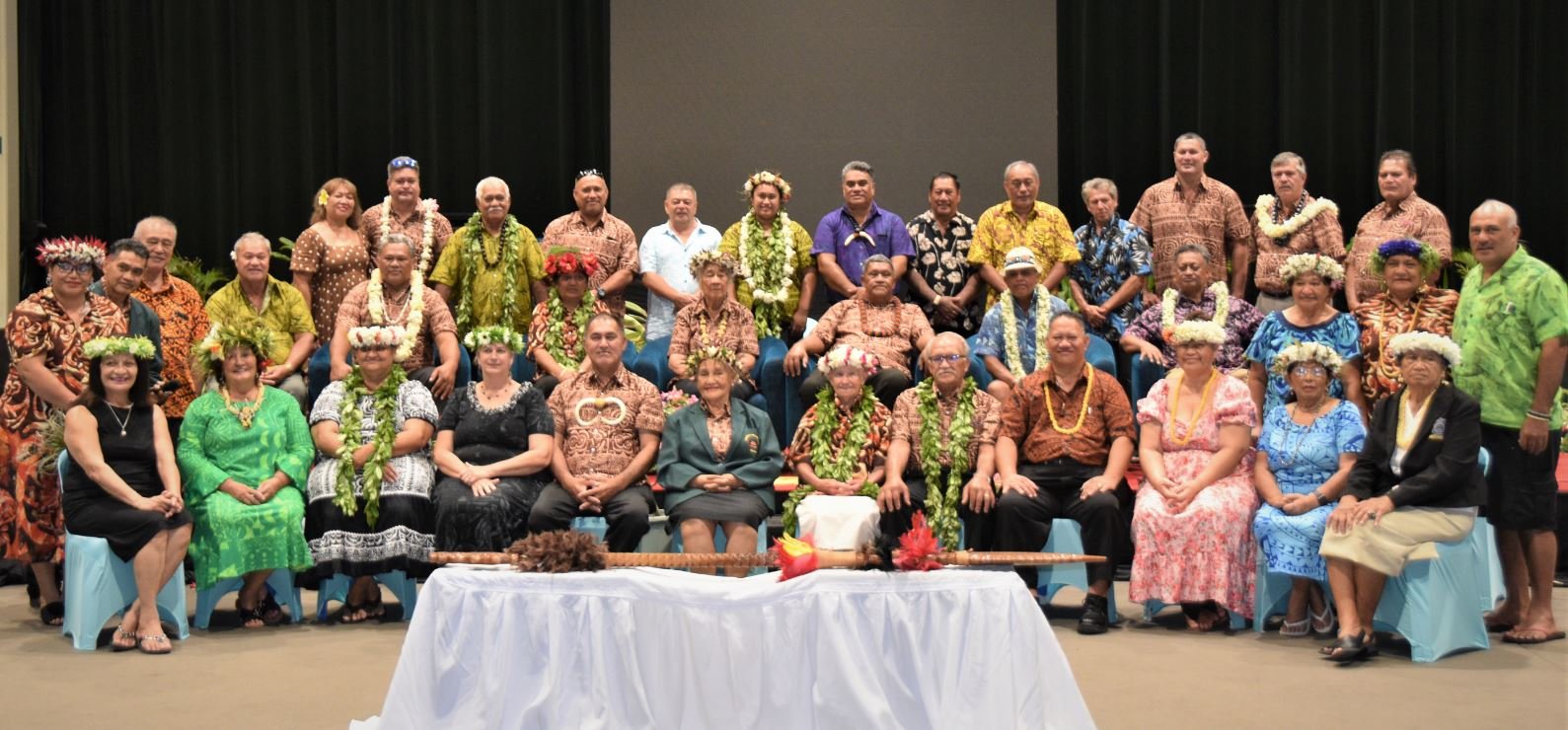 Group of people gathered in a room, dressed in traditional Hawaiian attire, some wearing flower leis and flower crowns, posing for a photo.