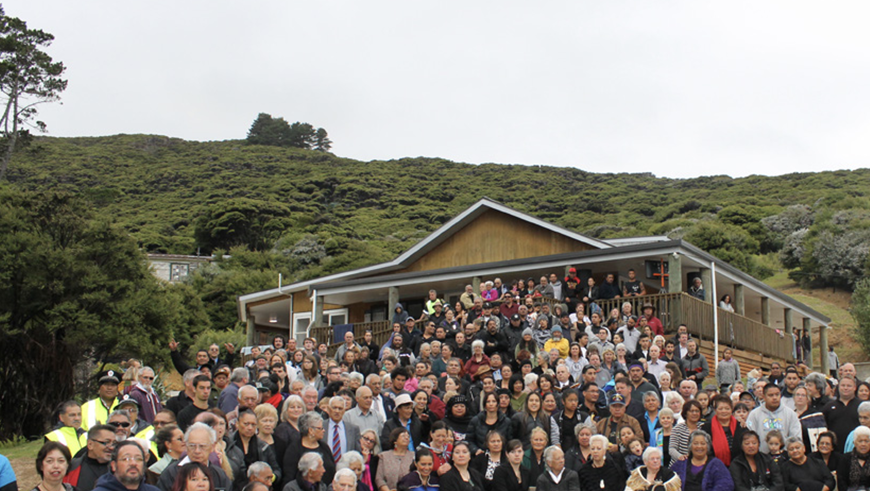 Large crowd of people gathered outside a house on a hillside surrounded by greenery.