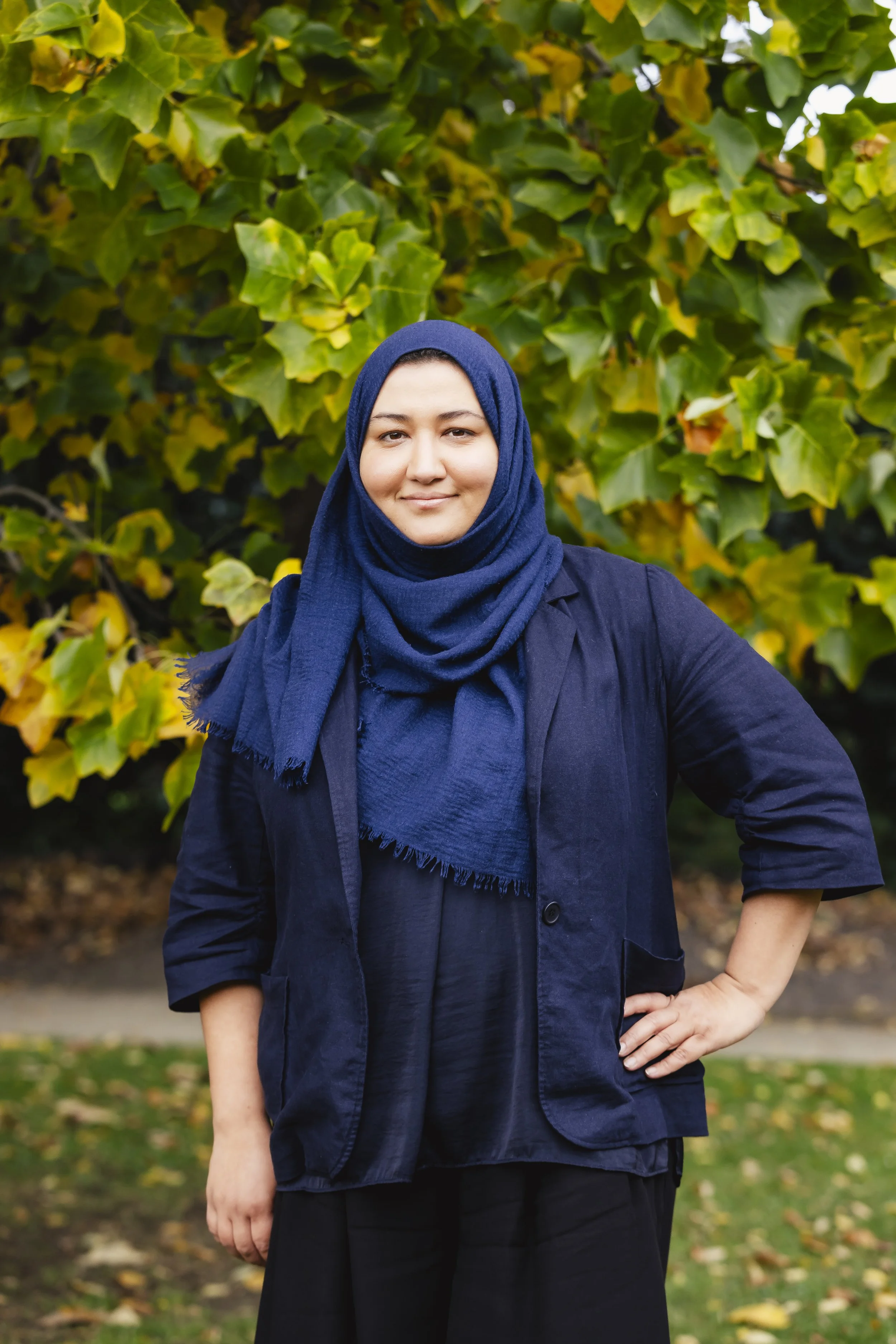 Woman wearing a blue hijab, dark blazer, and black pants standing outdoors with green and yellow foliage in the background.